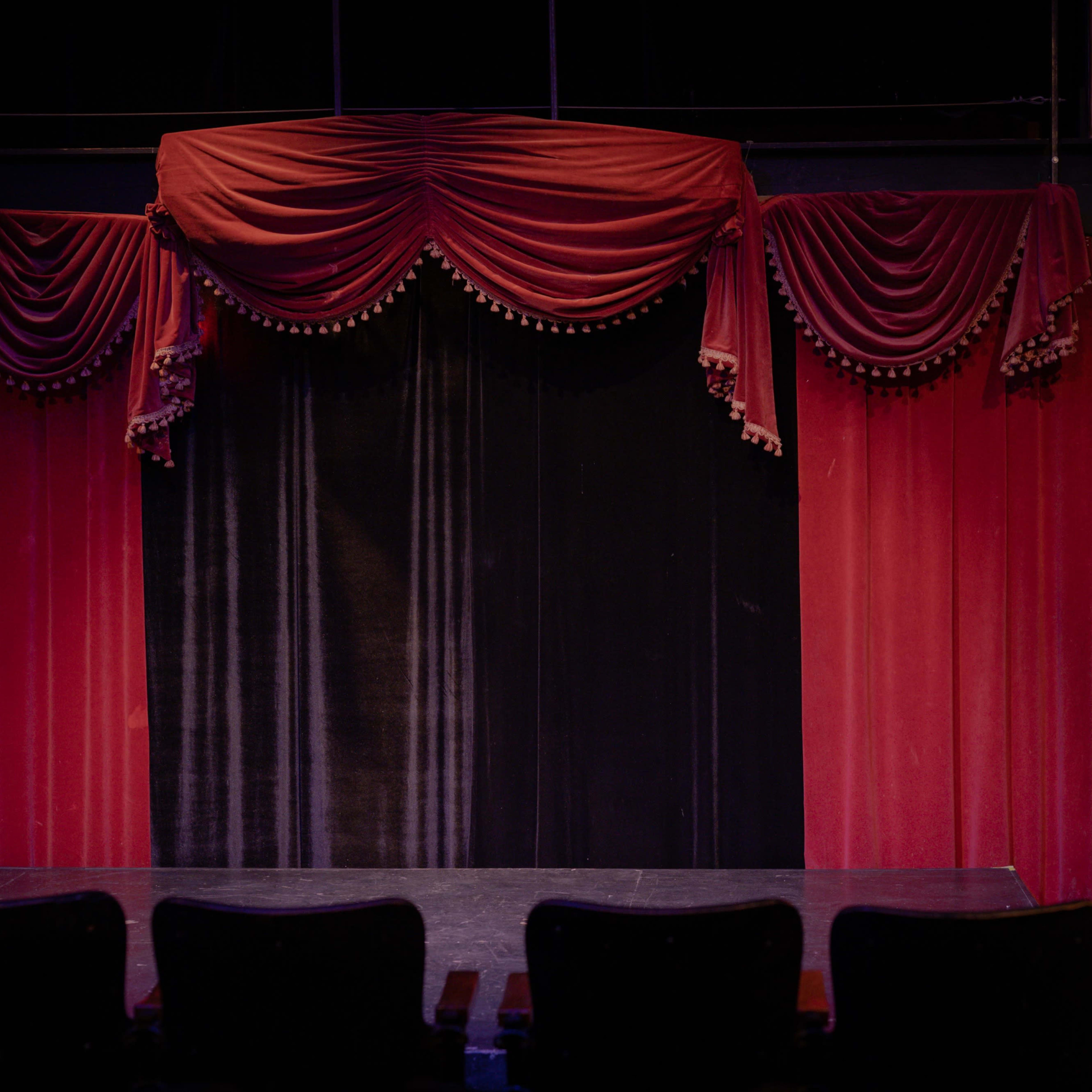A theater stage draped with red curtains, framed by ornate valances, and with empty black seats in front.