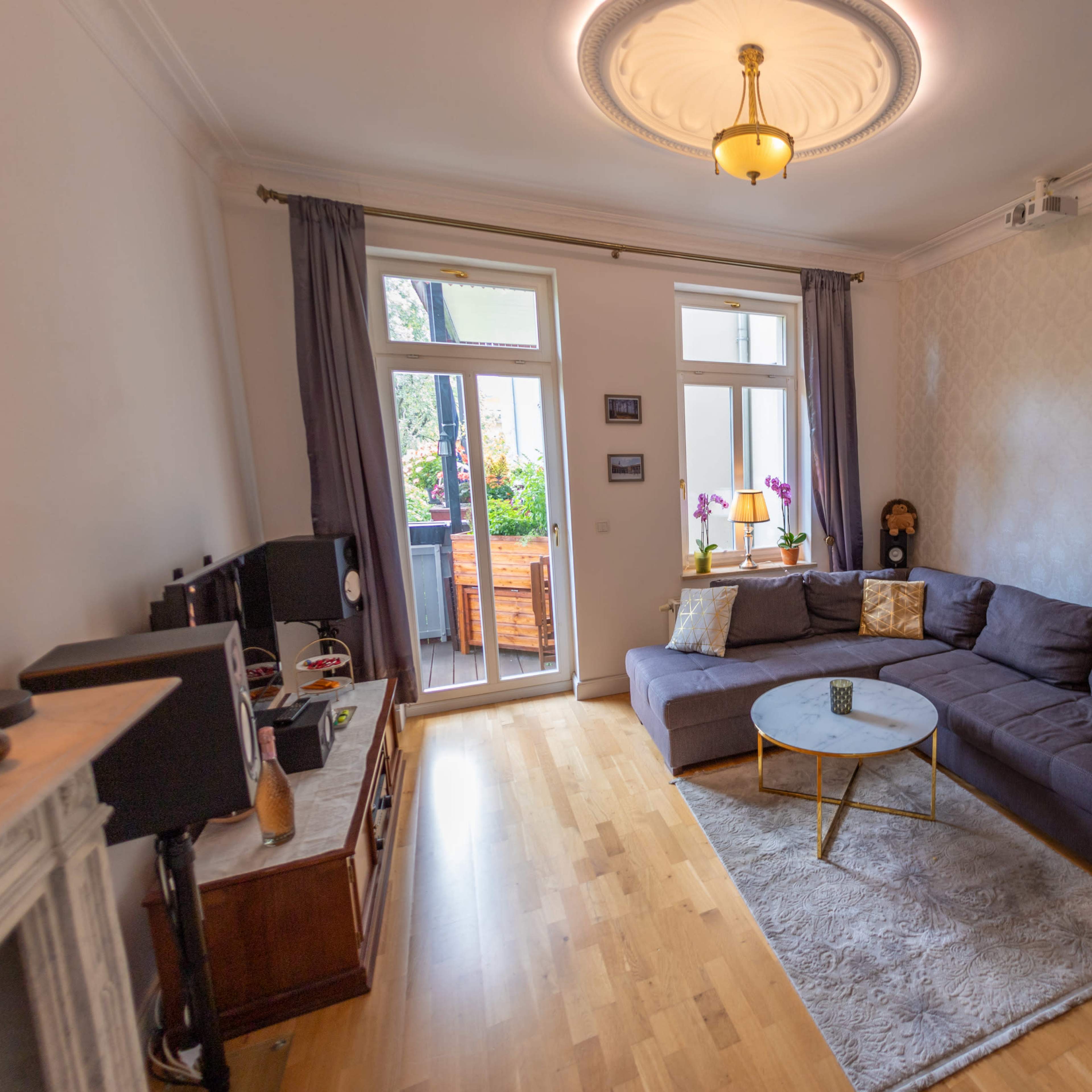 A living room features a dark sofa, a small round table, and large windows that open to a balcony, with light-colored walls and wooden flooring.