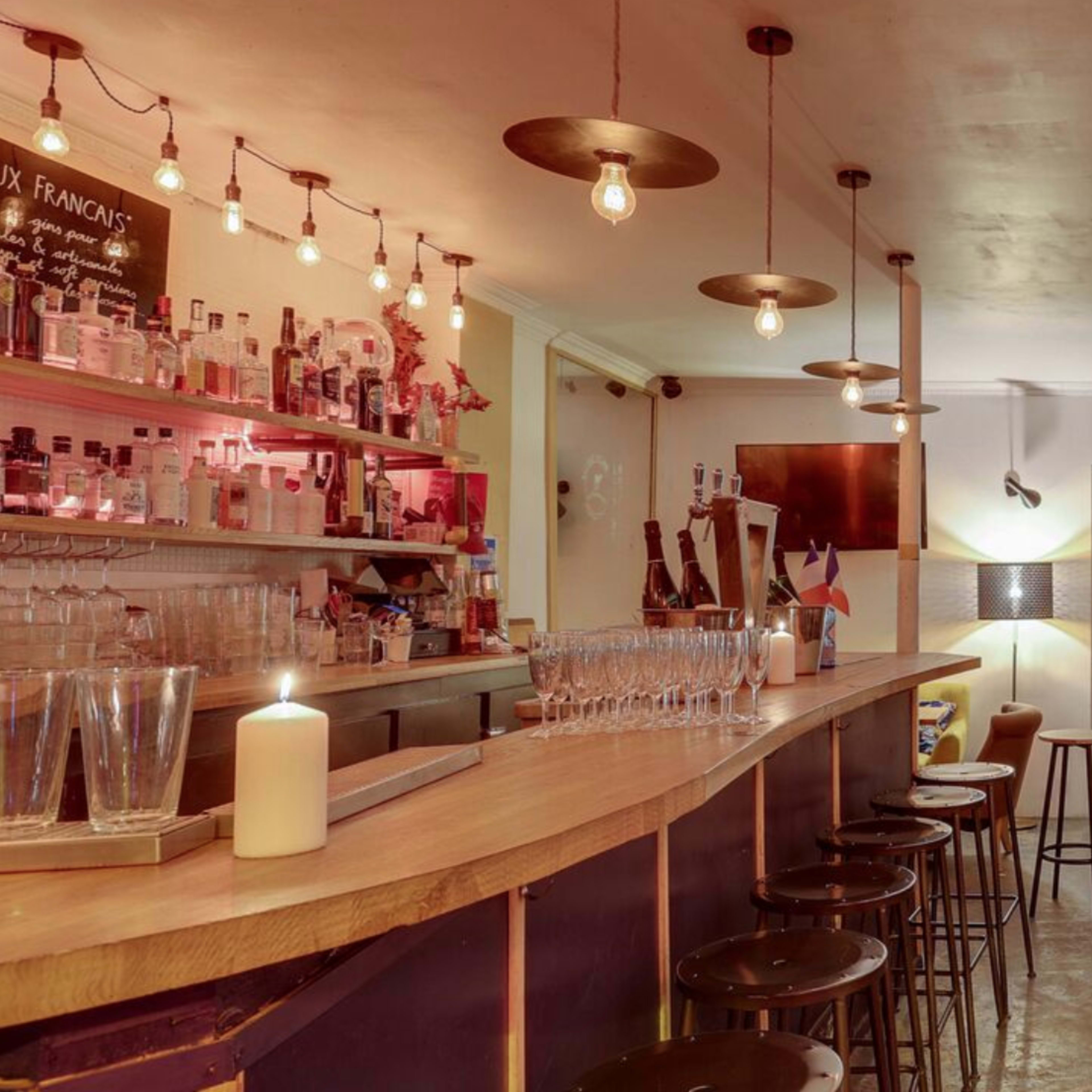 A dimly lit bar with a wooden counter, high stools, and a shelf of various alcoholic beverages.