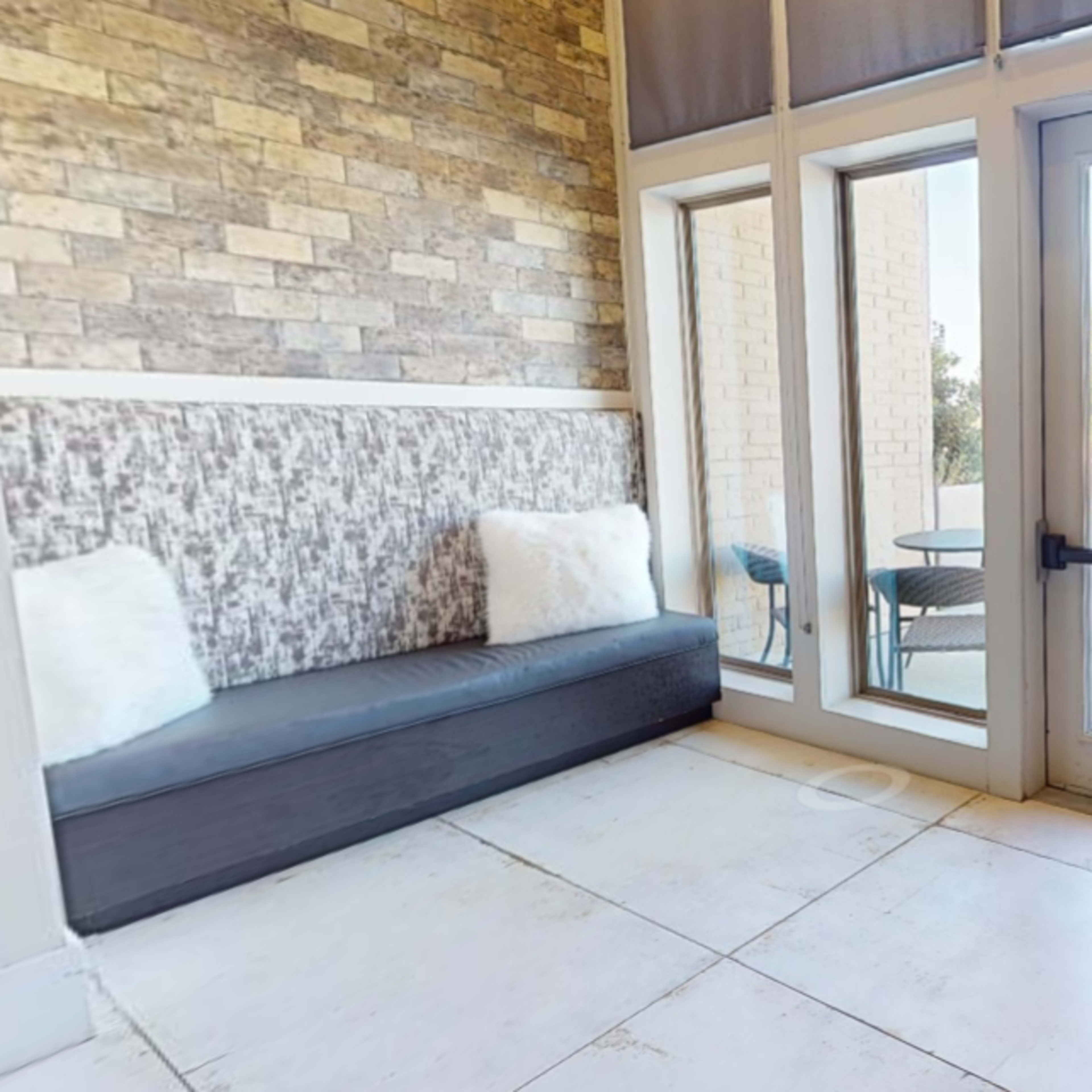 A cozy seating area with large windows, a wooden bench, and an armchair with decorative pillows, overlooking an outdoor space.