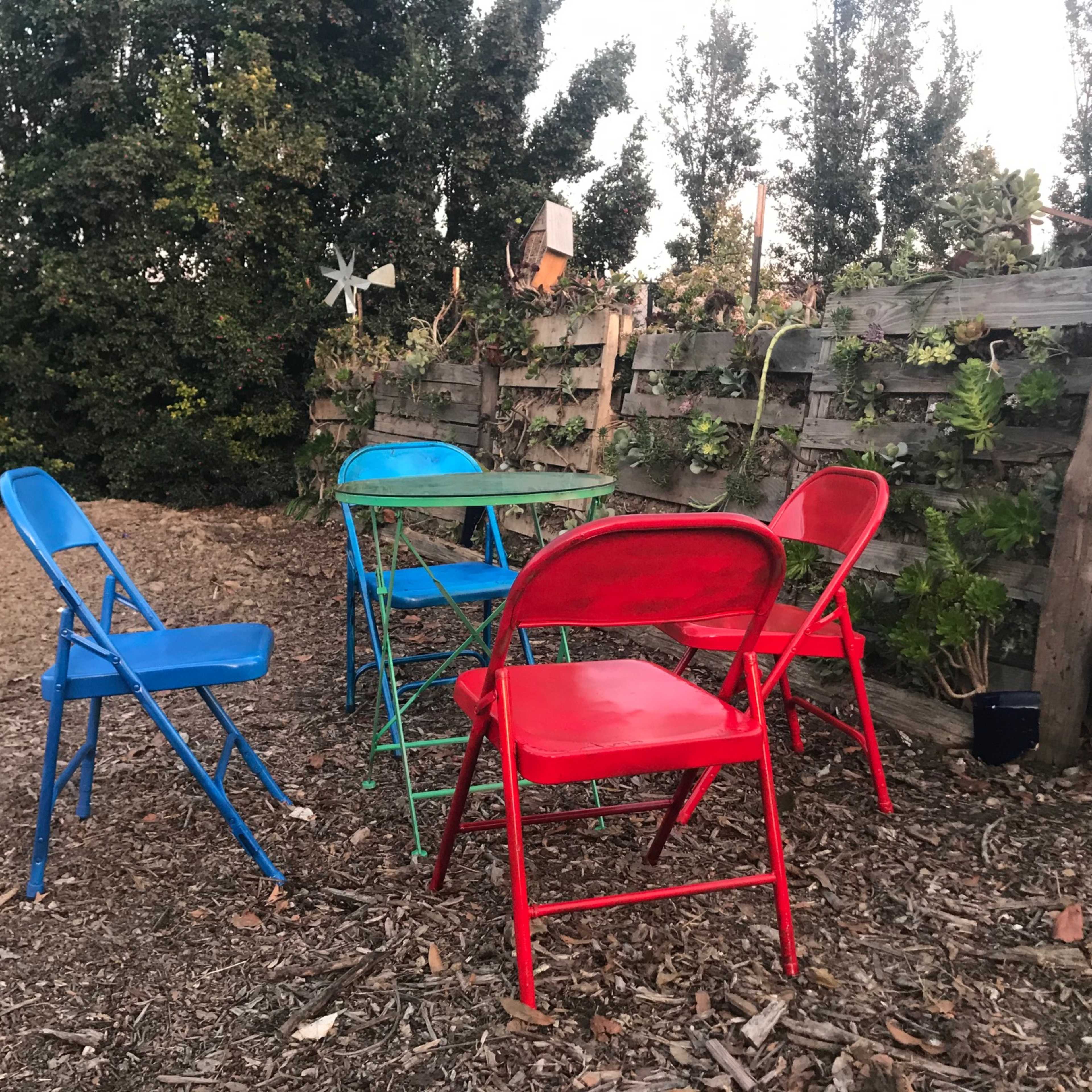 A set of colorful metal chairs surrounds a small, green table in a garden area with wooden fencing and plants.