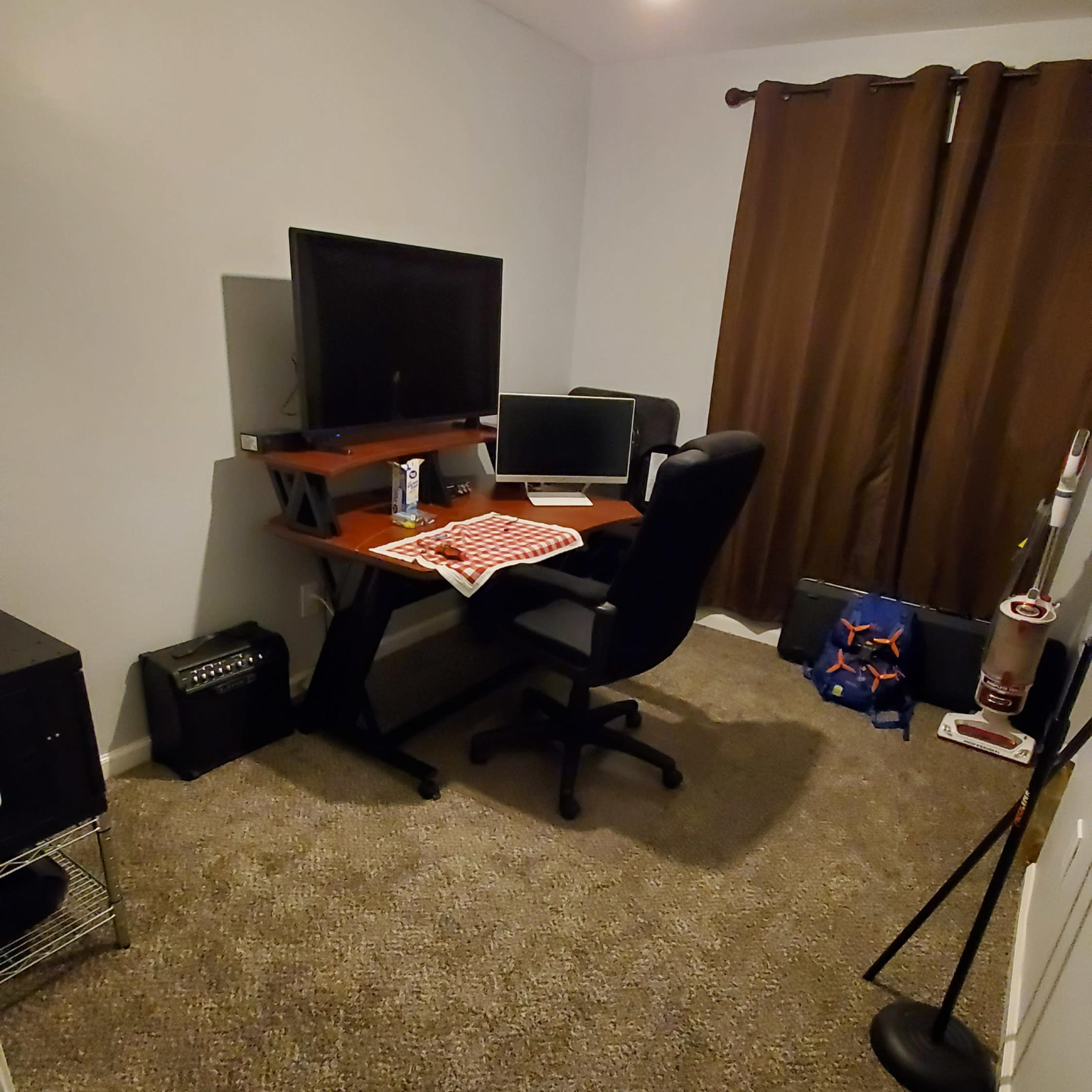 A small home office with a desk, two computer monitors, a black office chair, and a brown curtain covering a window.