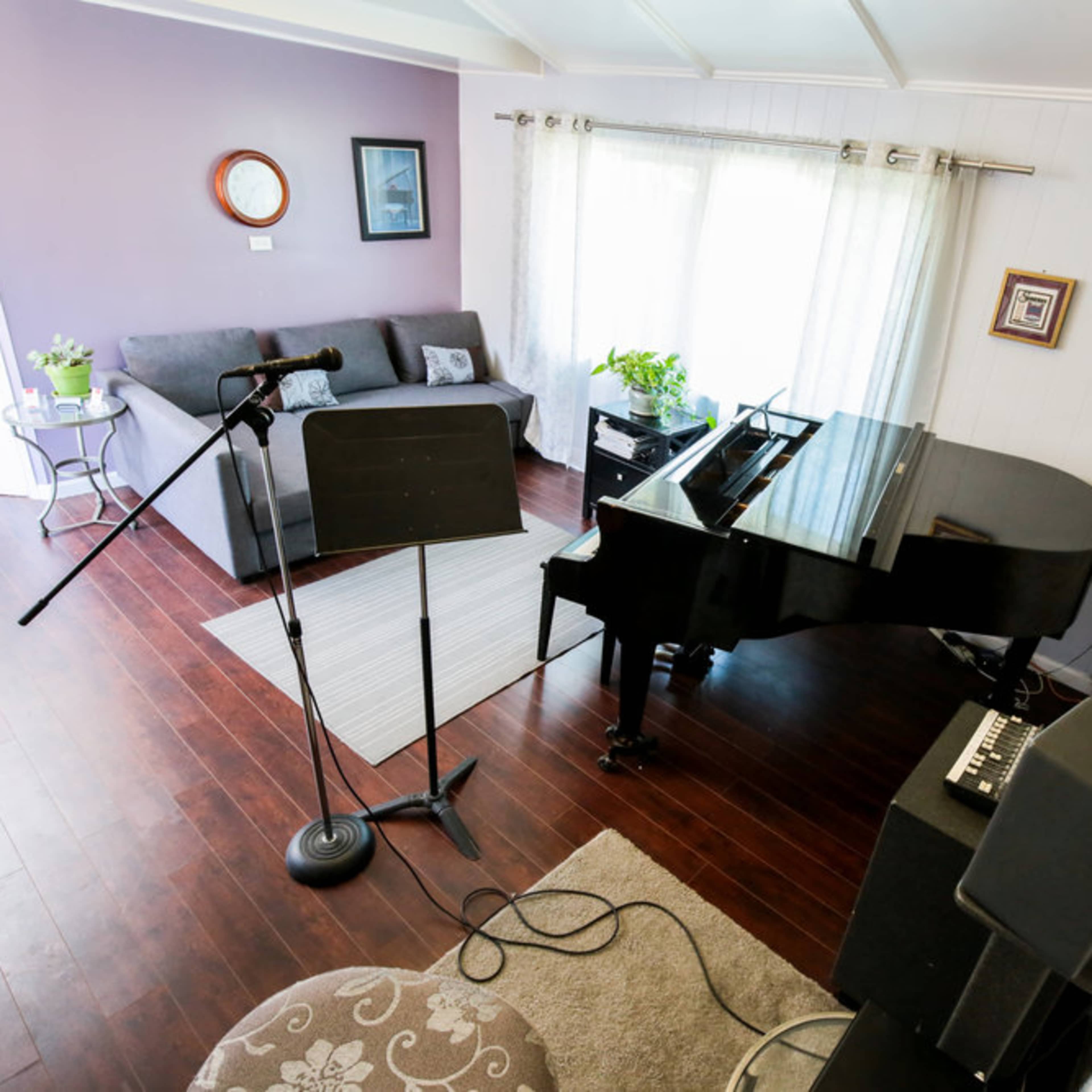 A living room features a grand piano, a microphone stand, a gray sofa, and several plants near a window.