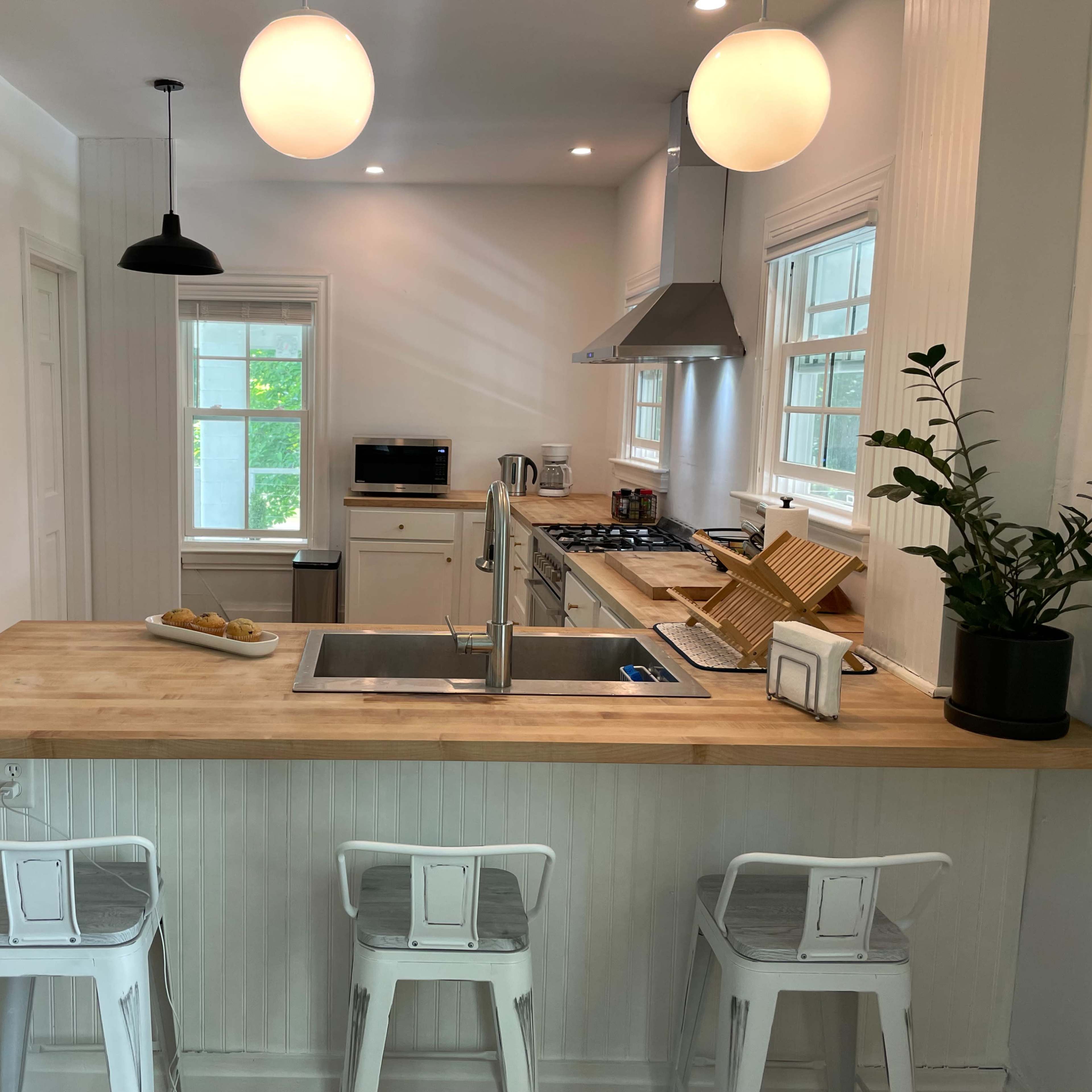 A modern kitchen features a wooden island with three stools, stainless steel appliances, and large windows letting in natural light.
