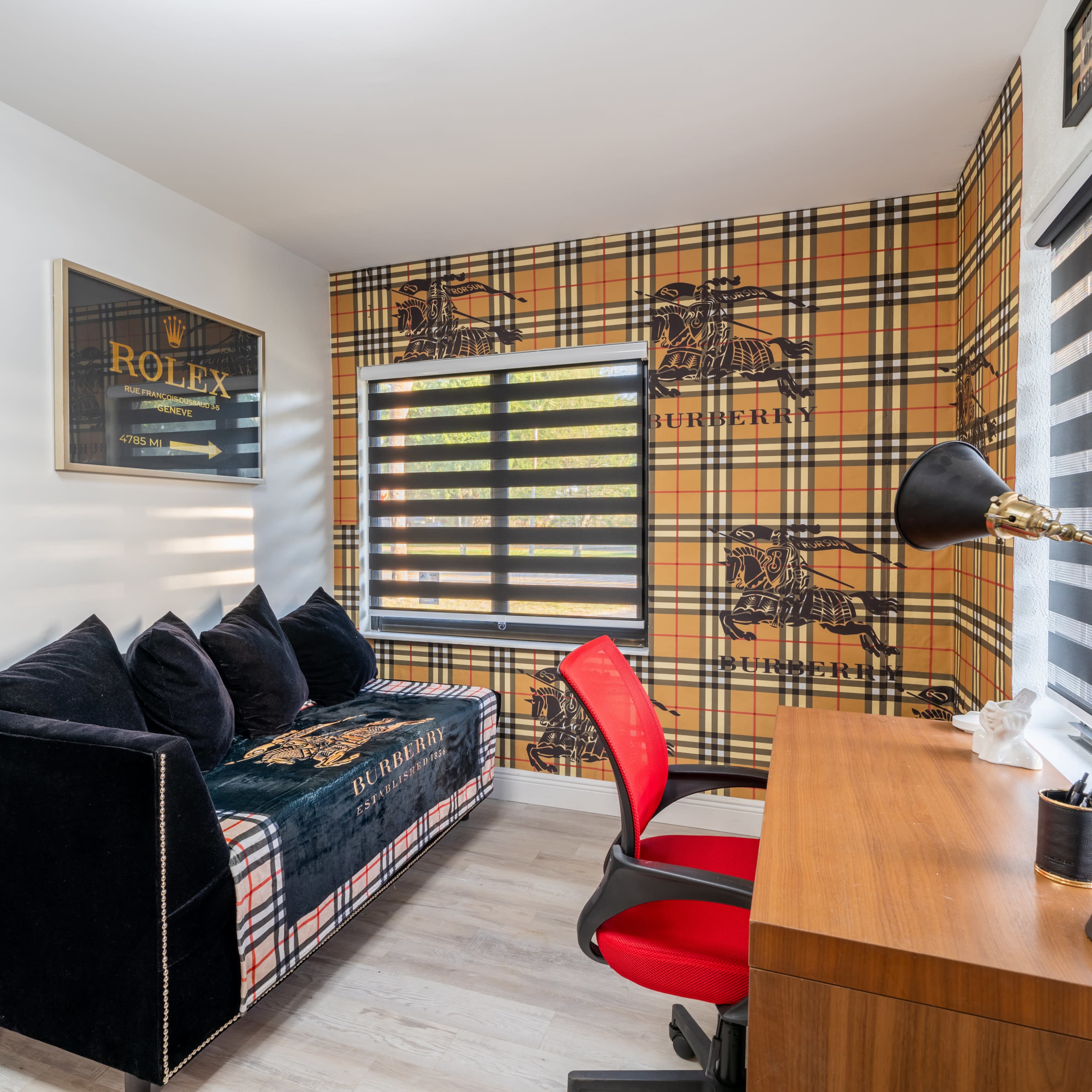A stylish office space featuring a plaid wallpaper, a black couch, a wooden desk with a lamp, and a large window with striped blinds.