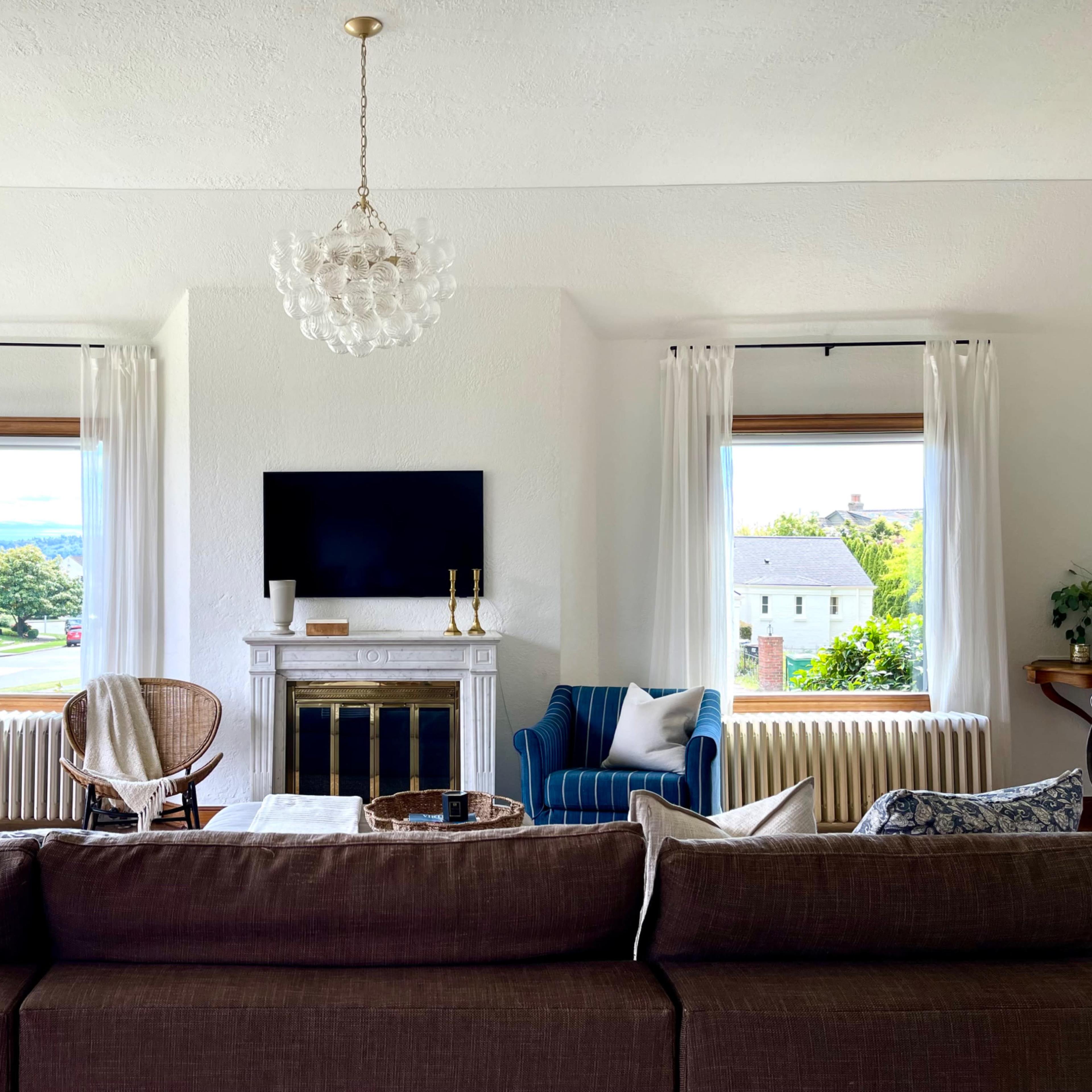 A living room features a fireplace with a television above it, two windows with curtains, a sofa, a blue armchair, and a decorative table in front.