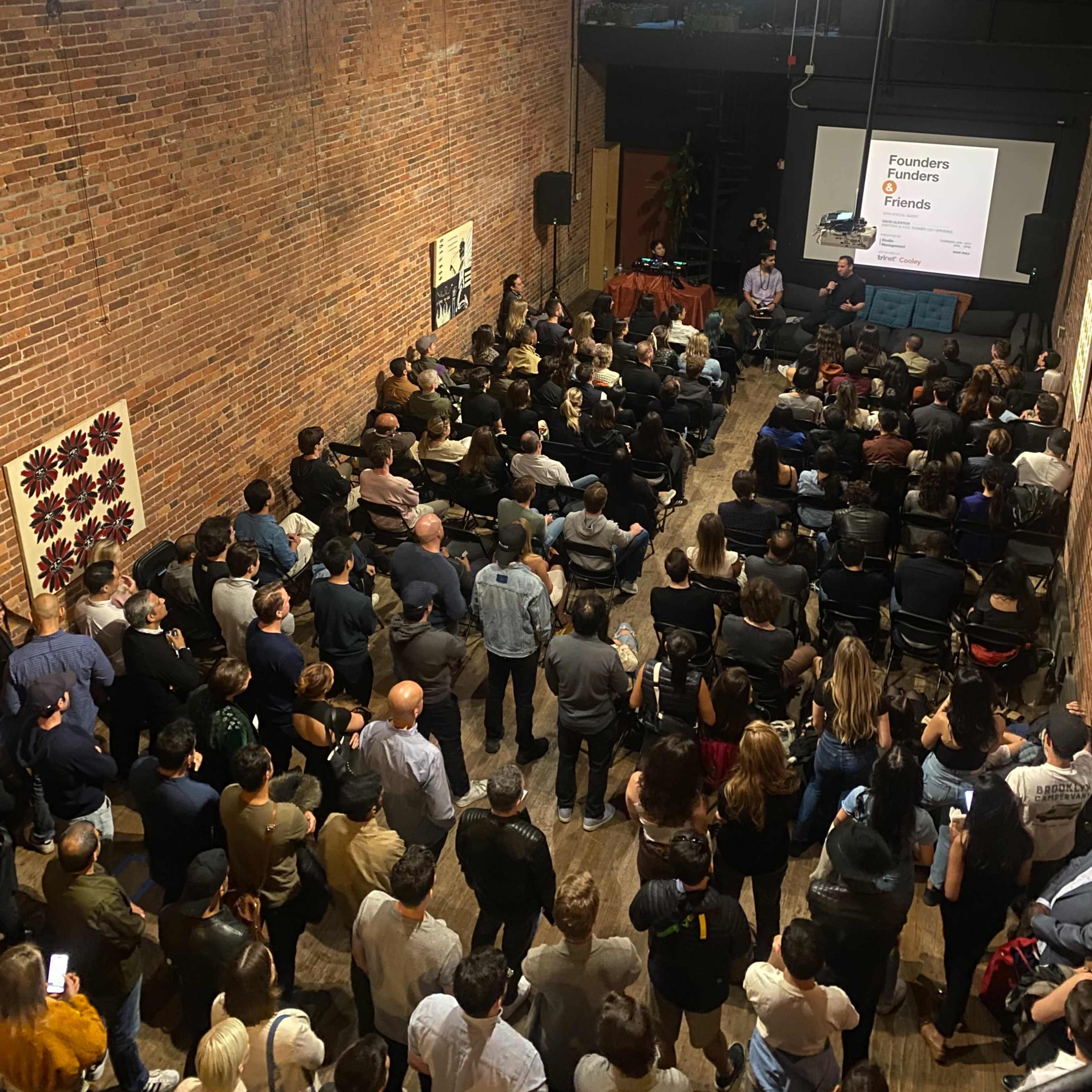 A large audience is seated in a brick-walled venue, attentively watching a presentation on a screen at the front.