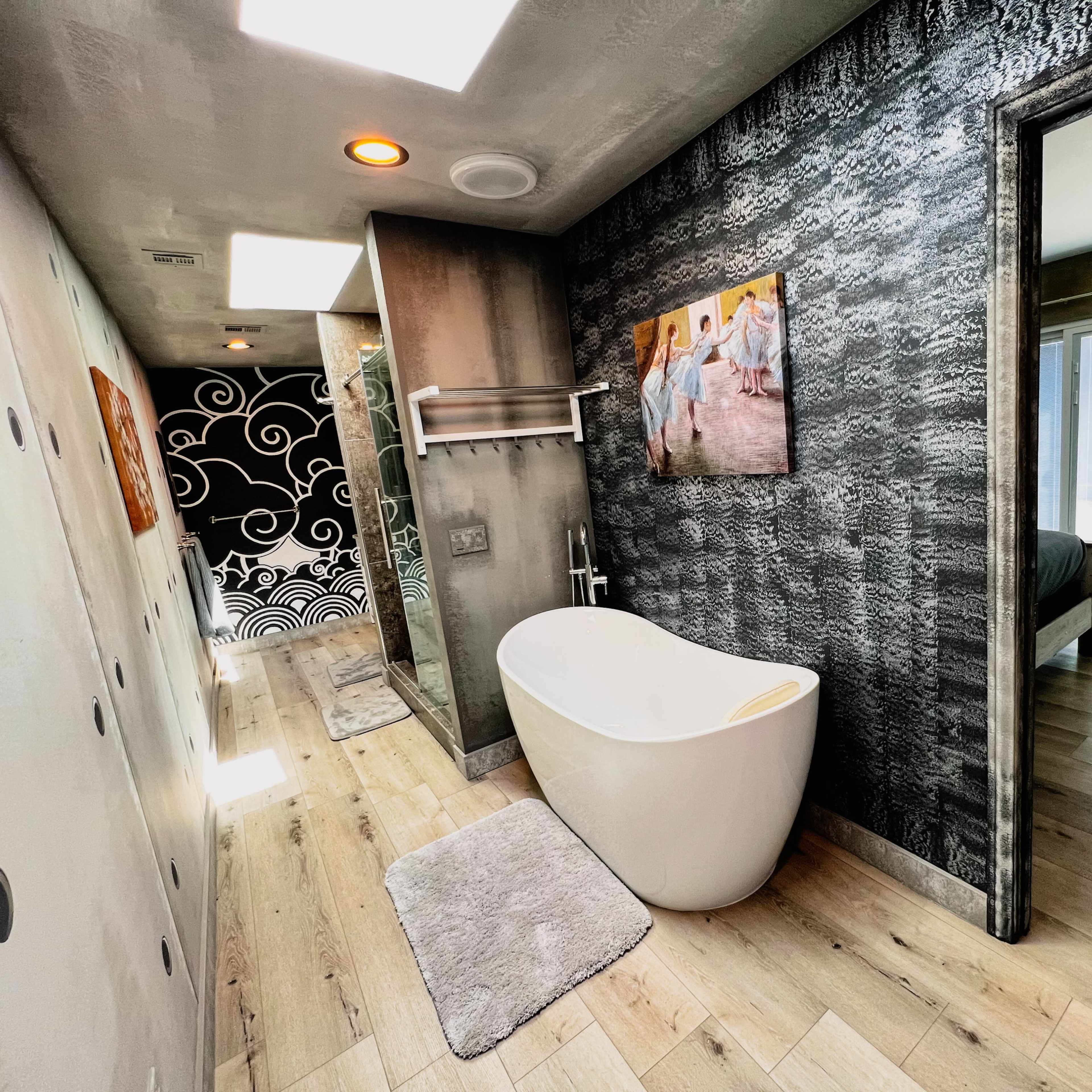 A modern bathroom with a freestanding white tub, wooden flooring, and a textured wall featuring a framed painting.