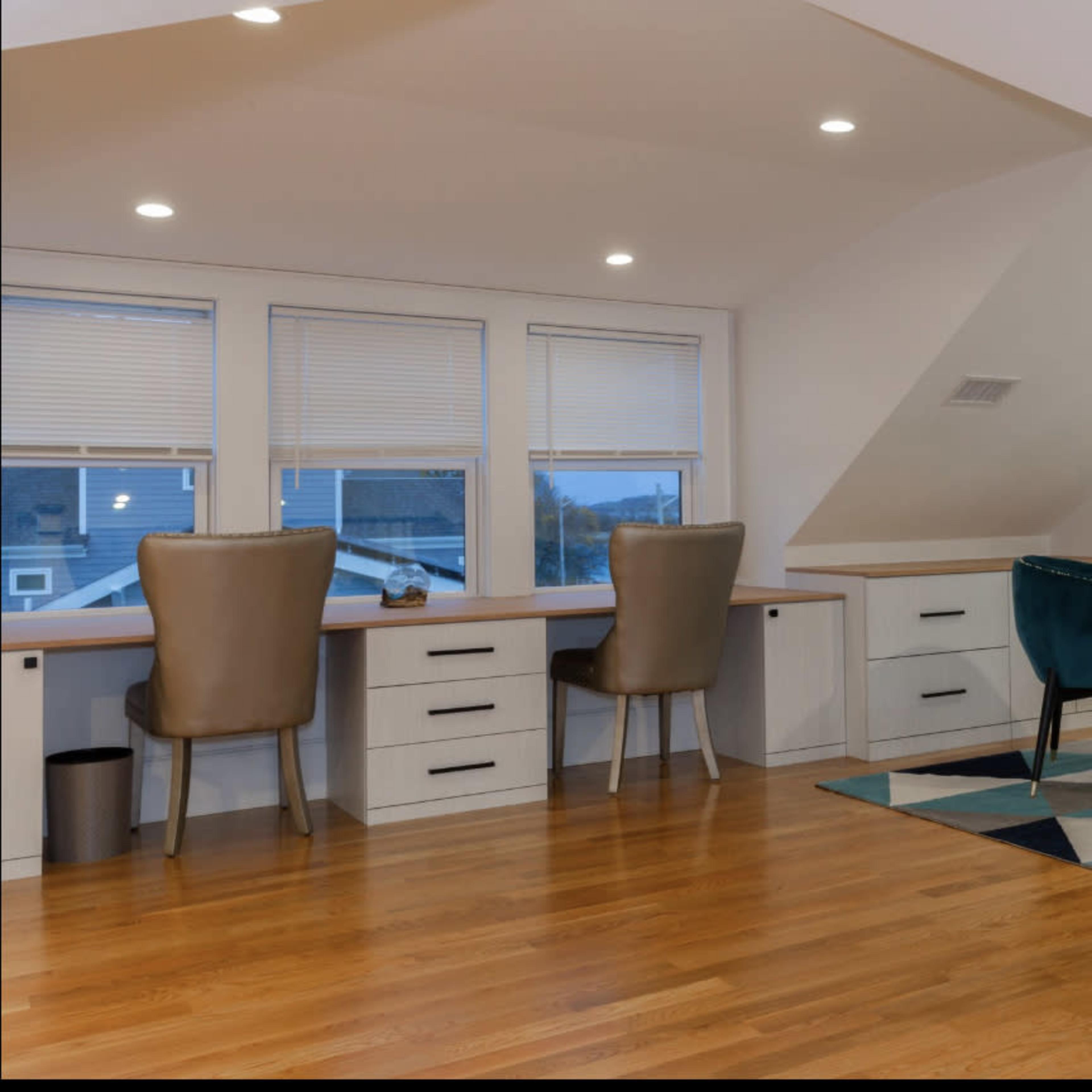 A modern, well-lit attic workspace featuring a wooden desk area with two chairs and a seating area with two teal chairs, situated near a large window.