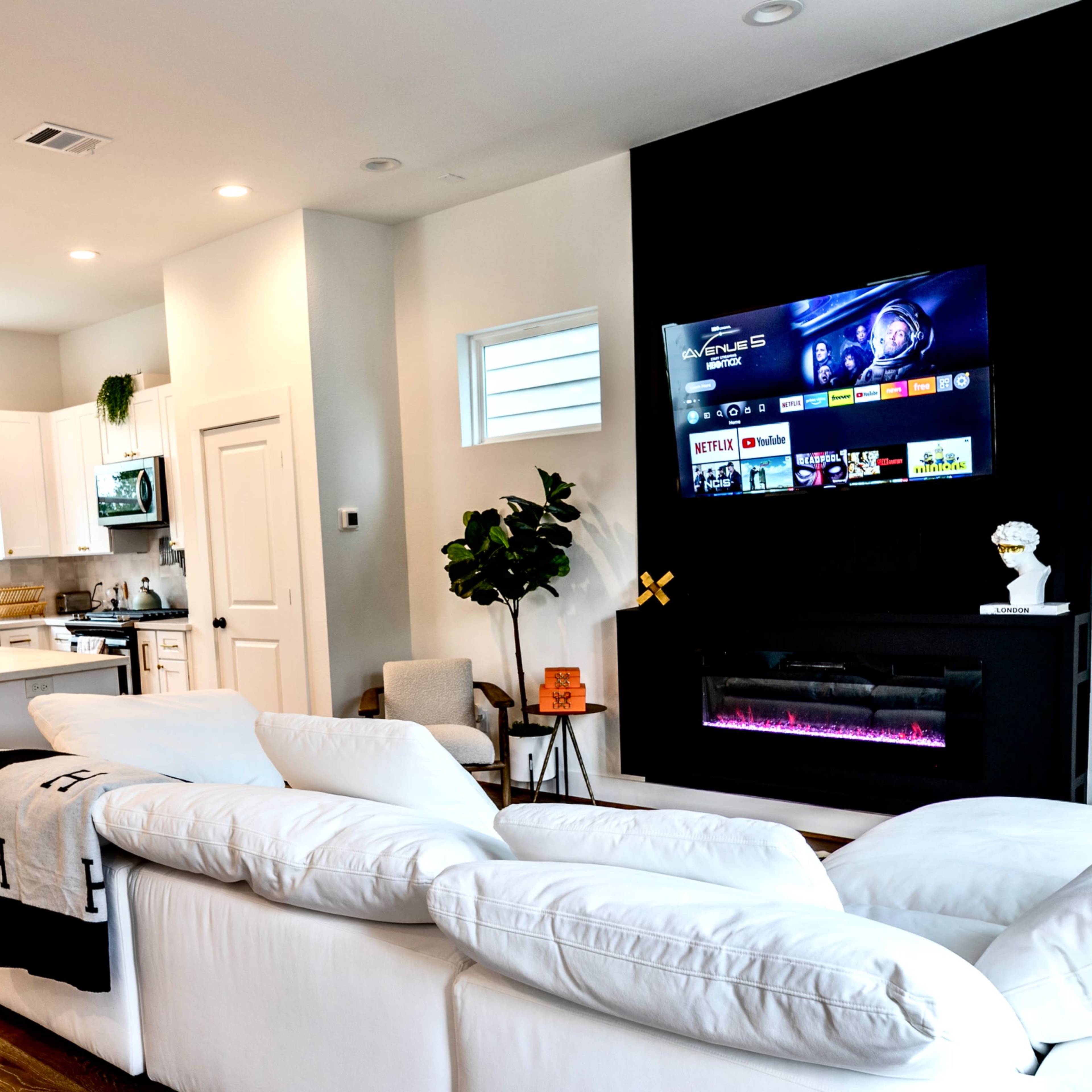 A modern living room features a white sectional sofa facing a wall-mounted TV above an electric fireplace, with a kitchen and dining area visible in the background.