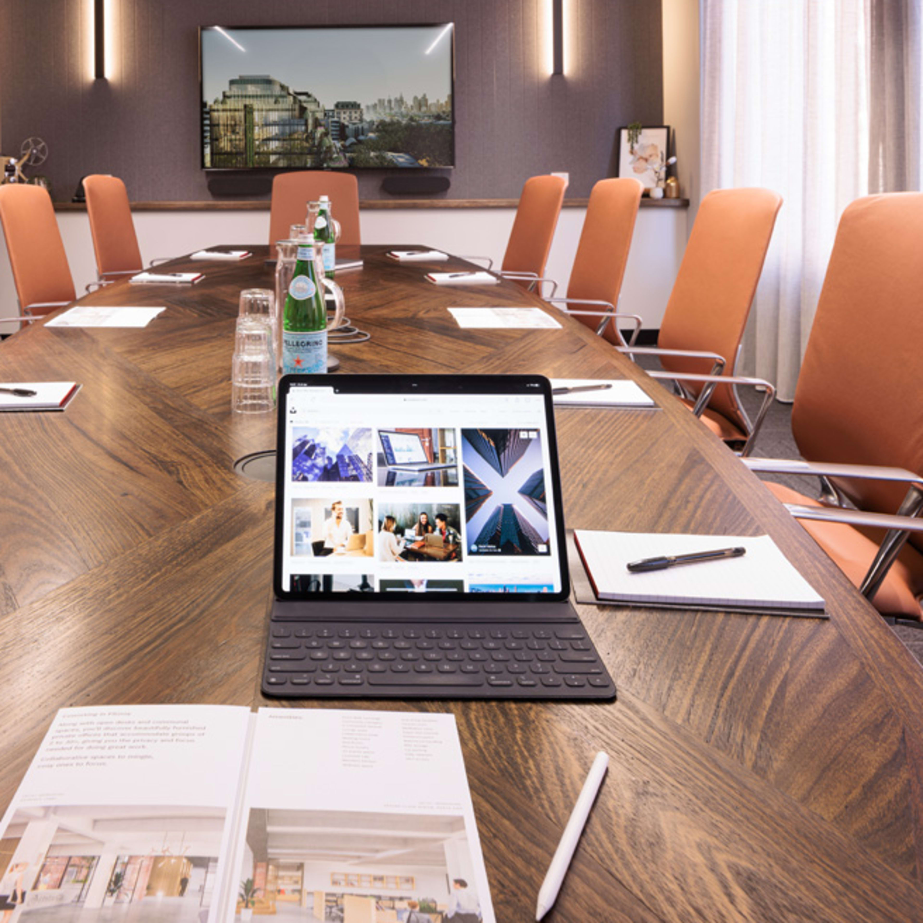 A modern conference room features an elongated wooden table, equipped with stationery and an iPad displaying images, surrounded by orange chairs.