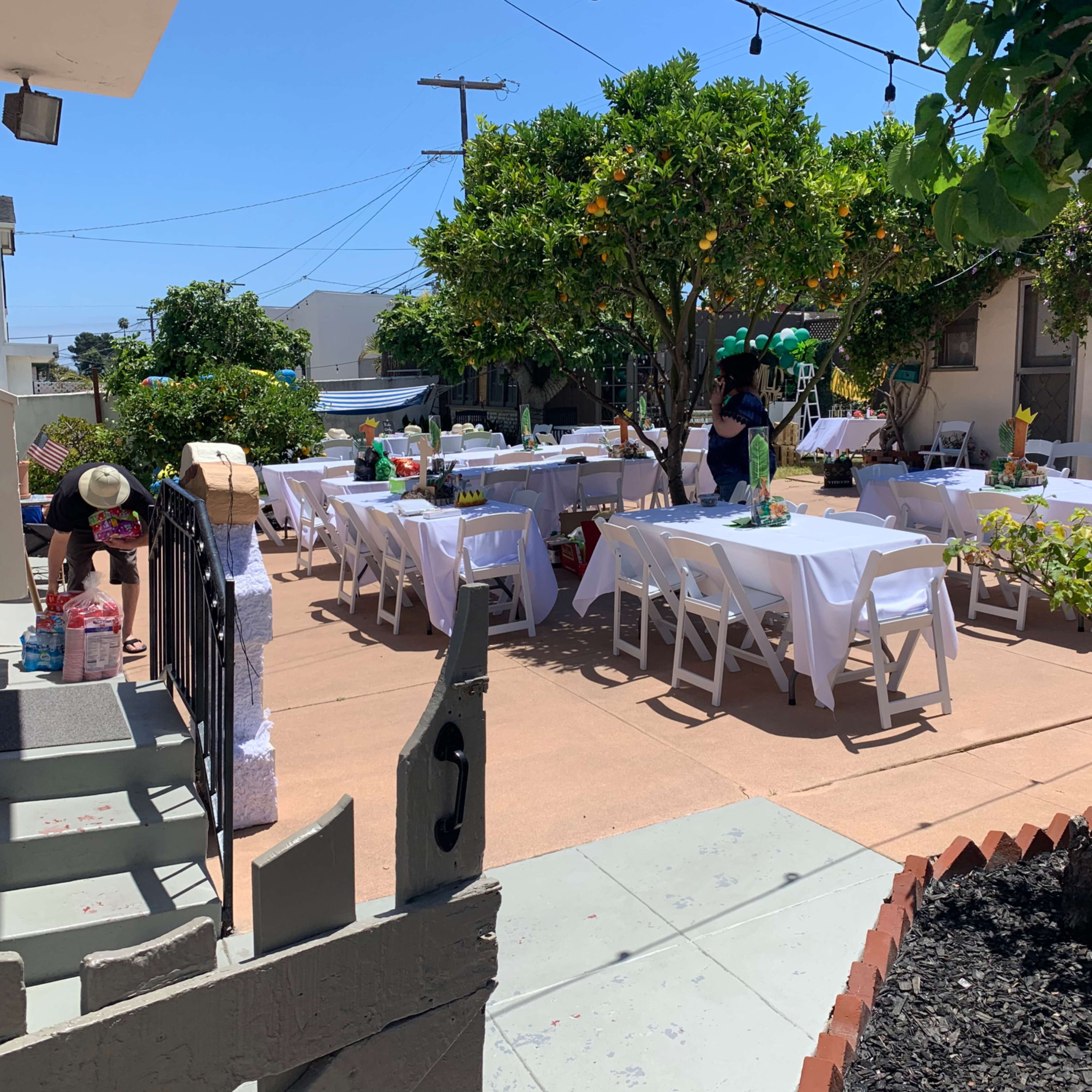 A garden party is set up with decorated tables and chairs in a courtyard surrounded by greenery and buildings.