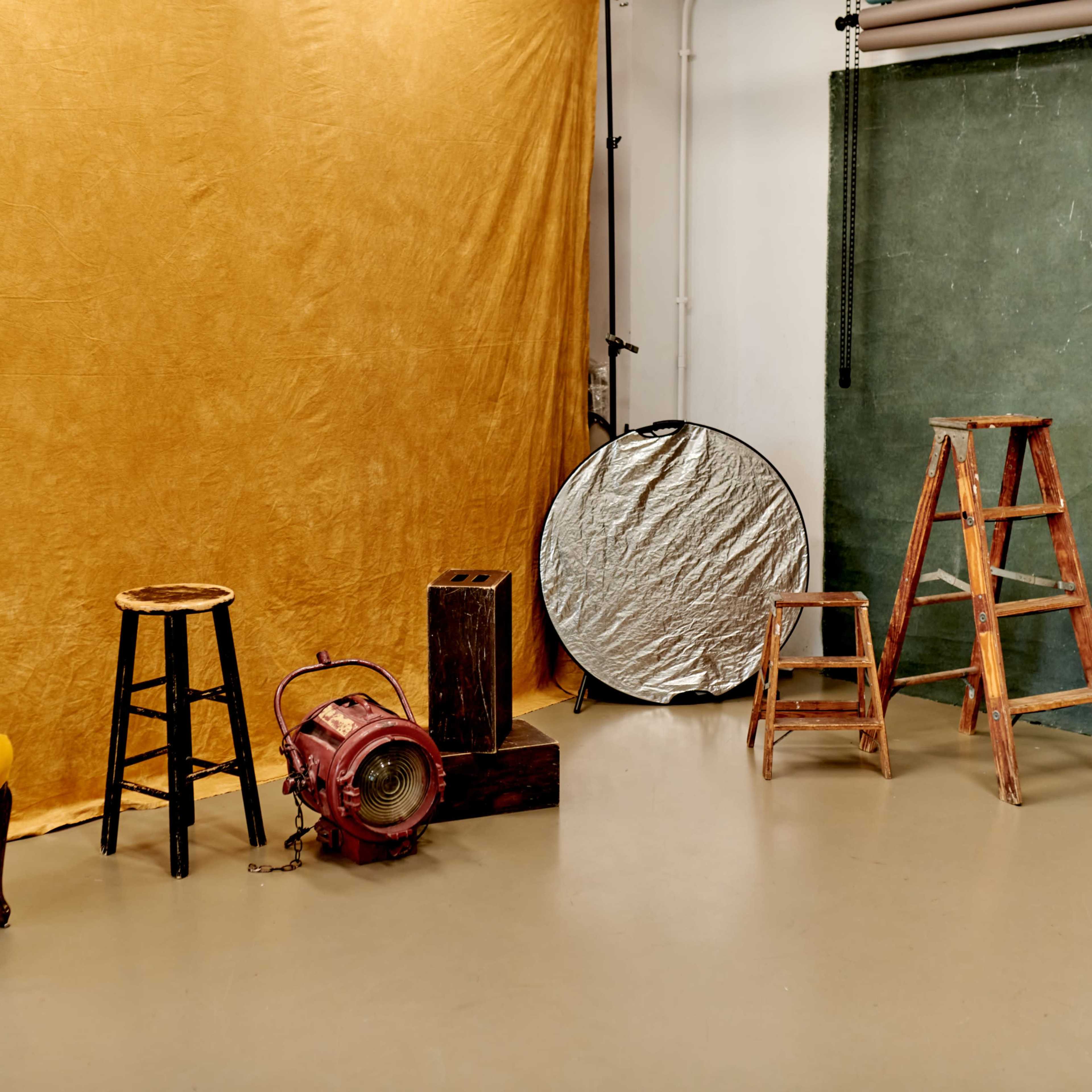 A photography studio setup with various props including a yellow chair, stools, a wooden ladder, and a reflector against a textured backdrop.