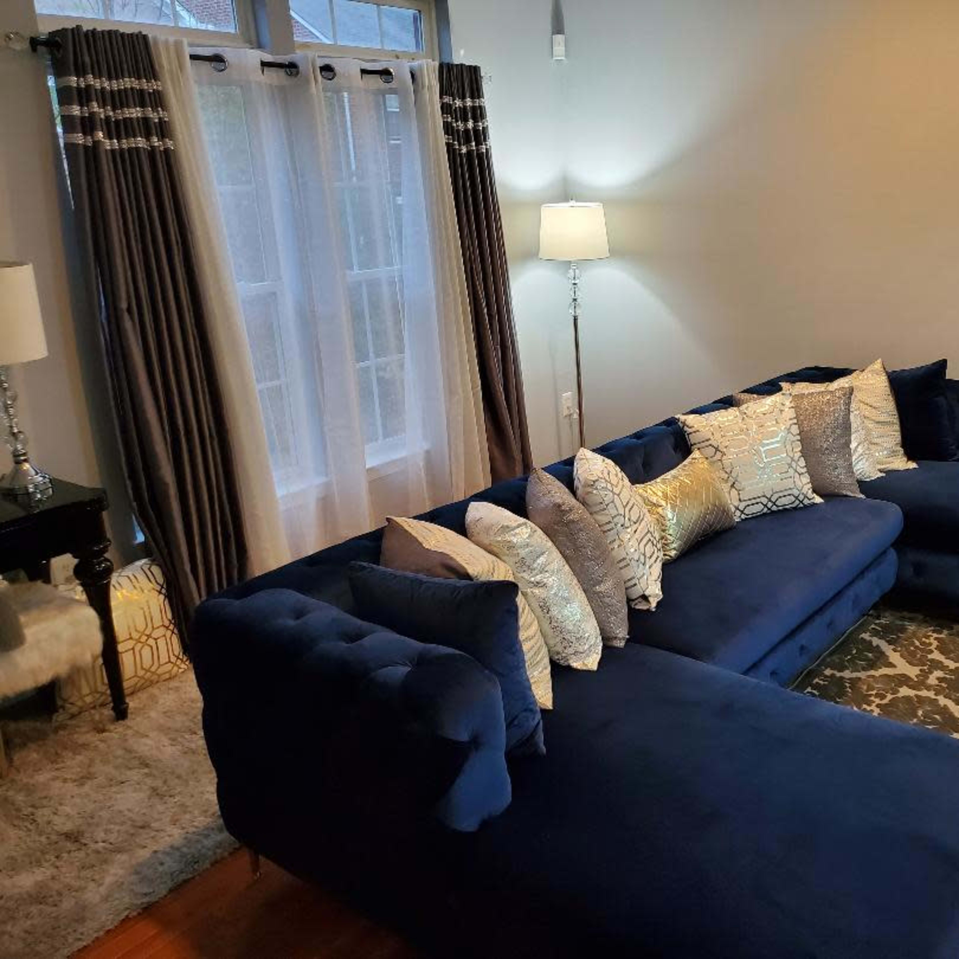 A modern living room features a navy blue sectional sofa adorned with decorative pillows, alongside a floor lamp and large windows draped with curtains.