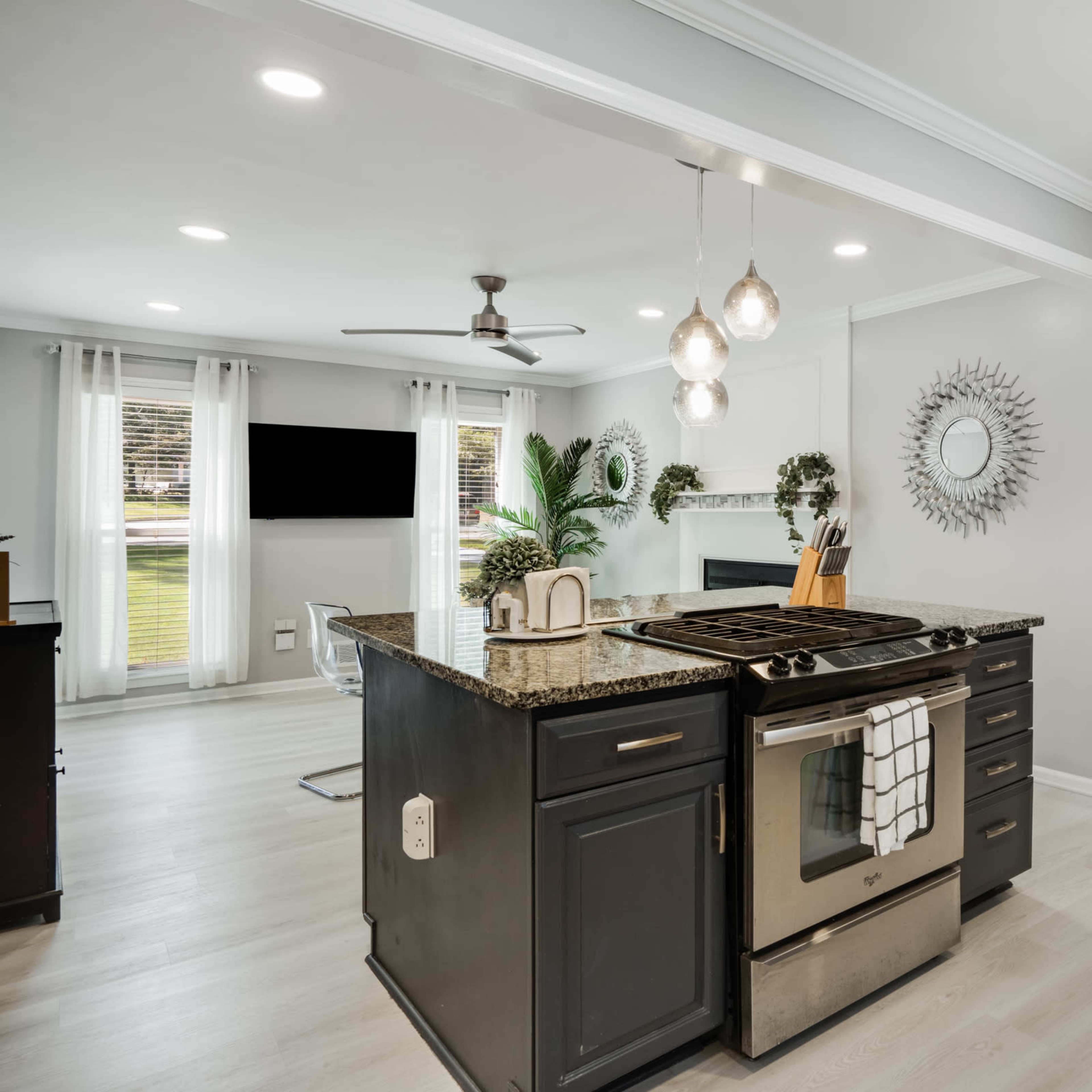 A modern kitchen with a central island, featuring a stove, granite countertops, and a view of an adjacent living area with natural light.