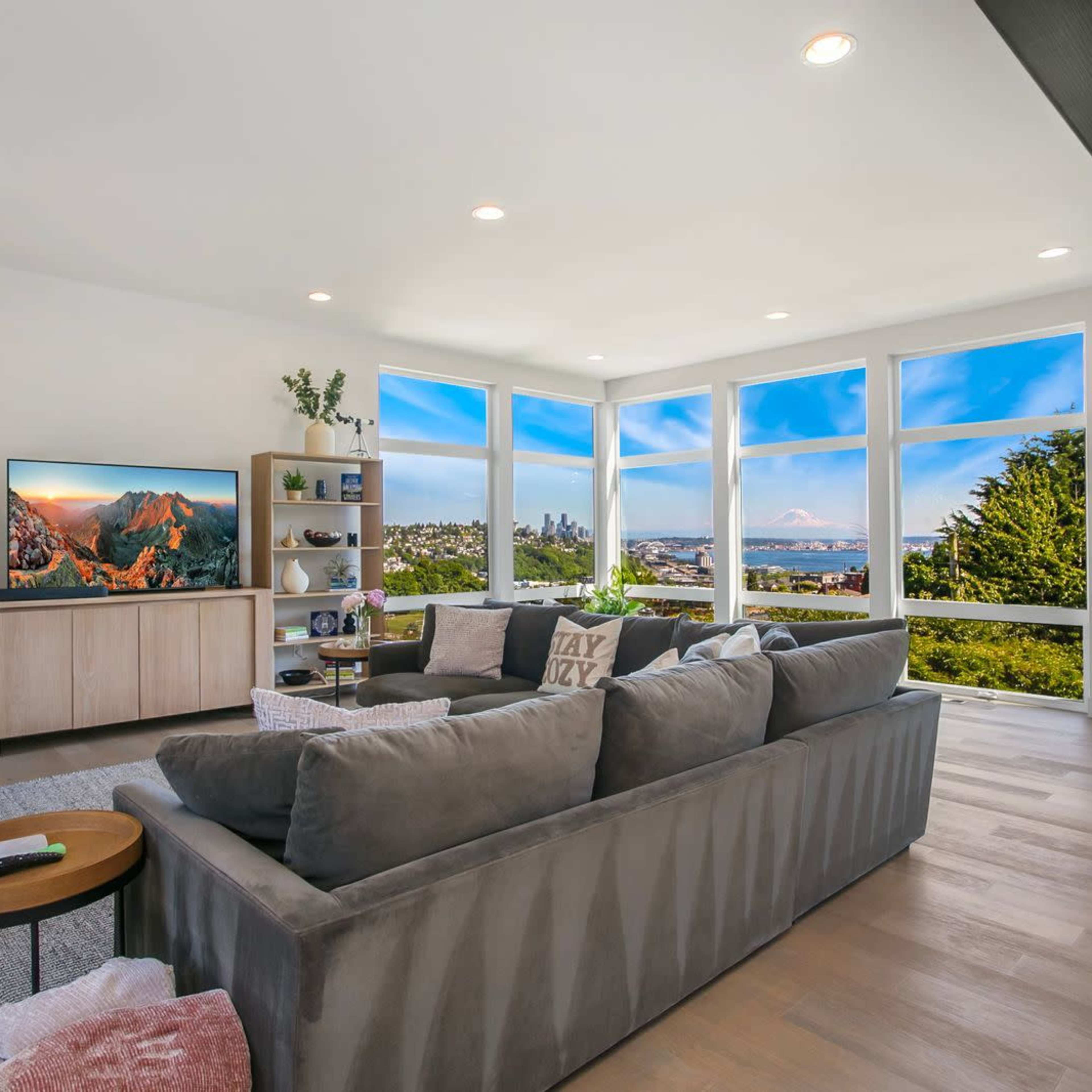 The living room features a large gray sofa facing a television, with floor-to-ceiling windows offering a view of the outdoors and surrounding landscape.