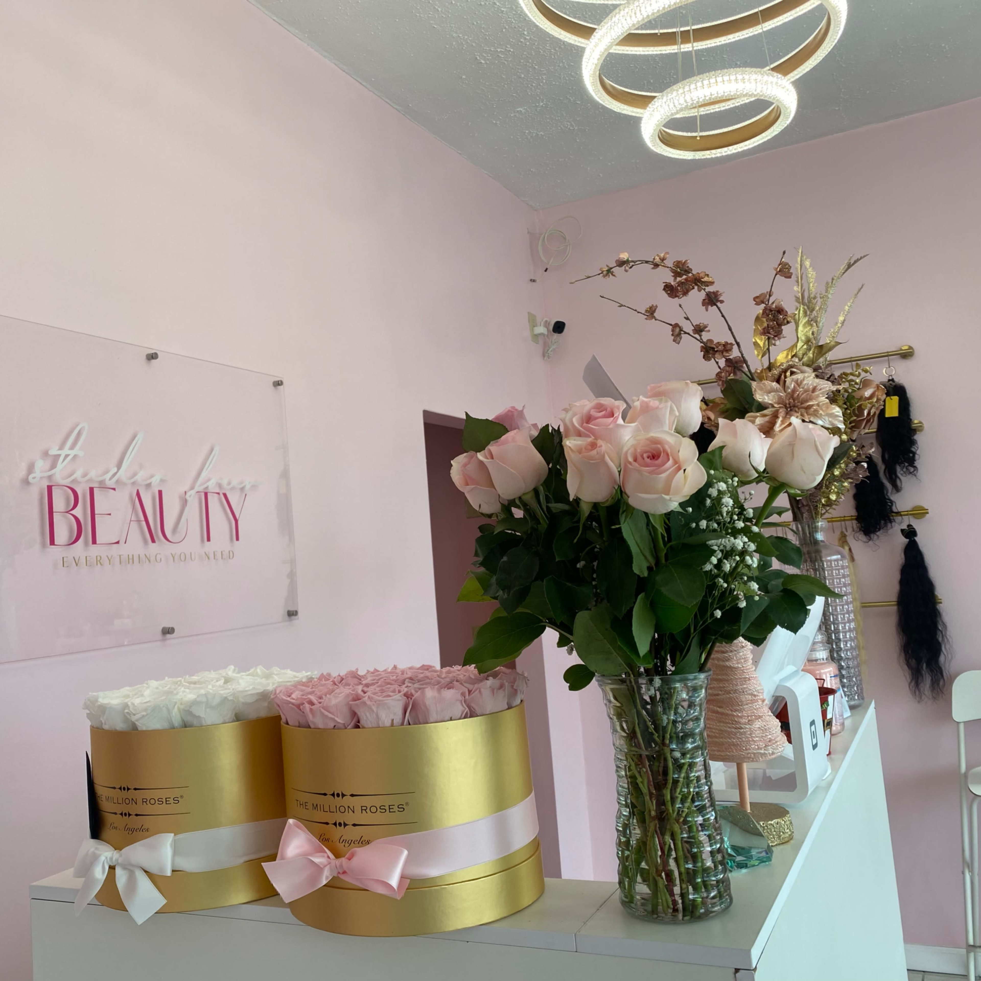 A beauty salon interior featuring a pink wall, a sign that reads "Salon of Beauty," and a counter adorned with two cylindrical flower arrangements and a vase of roses.