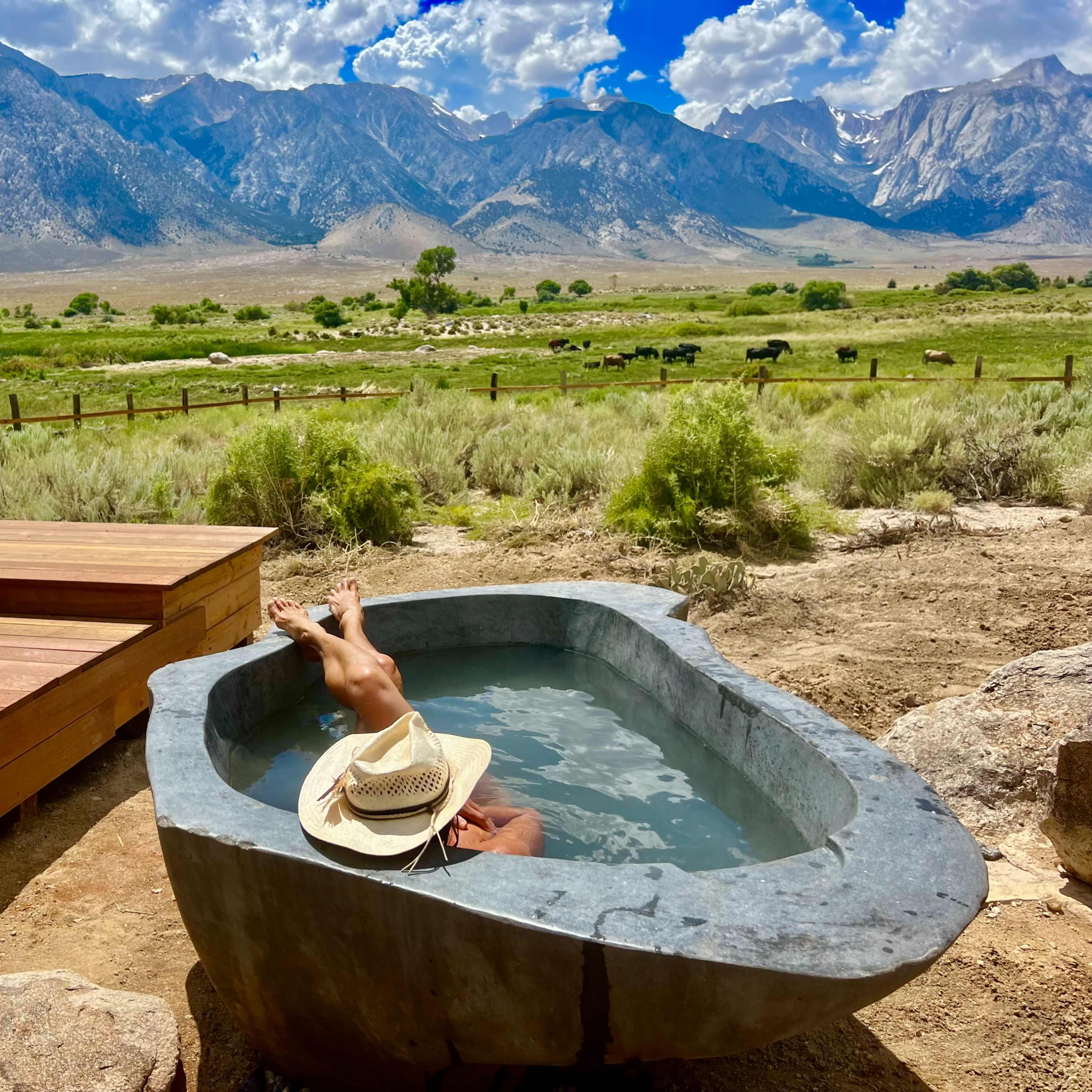A person relaxes in a stone bathtub surrounded by mountains, with a wooden deck and a statue in the background.