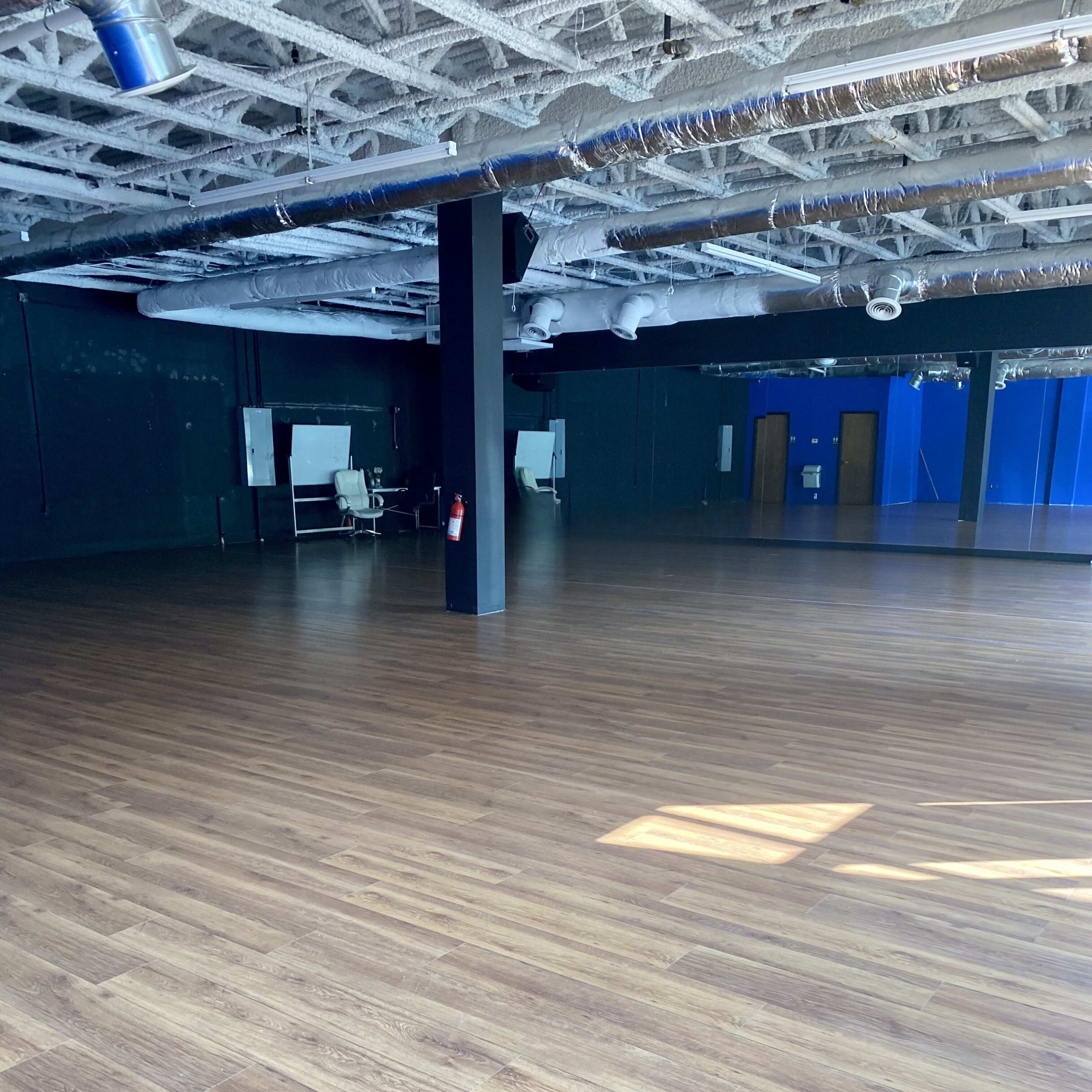 A spacious empty room with wooden flooring and black walls, featuring a few pieces of furniture in the background.