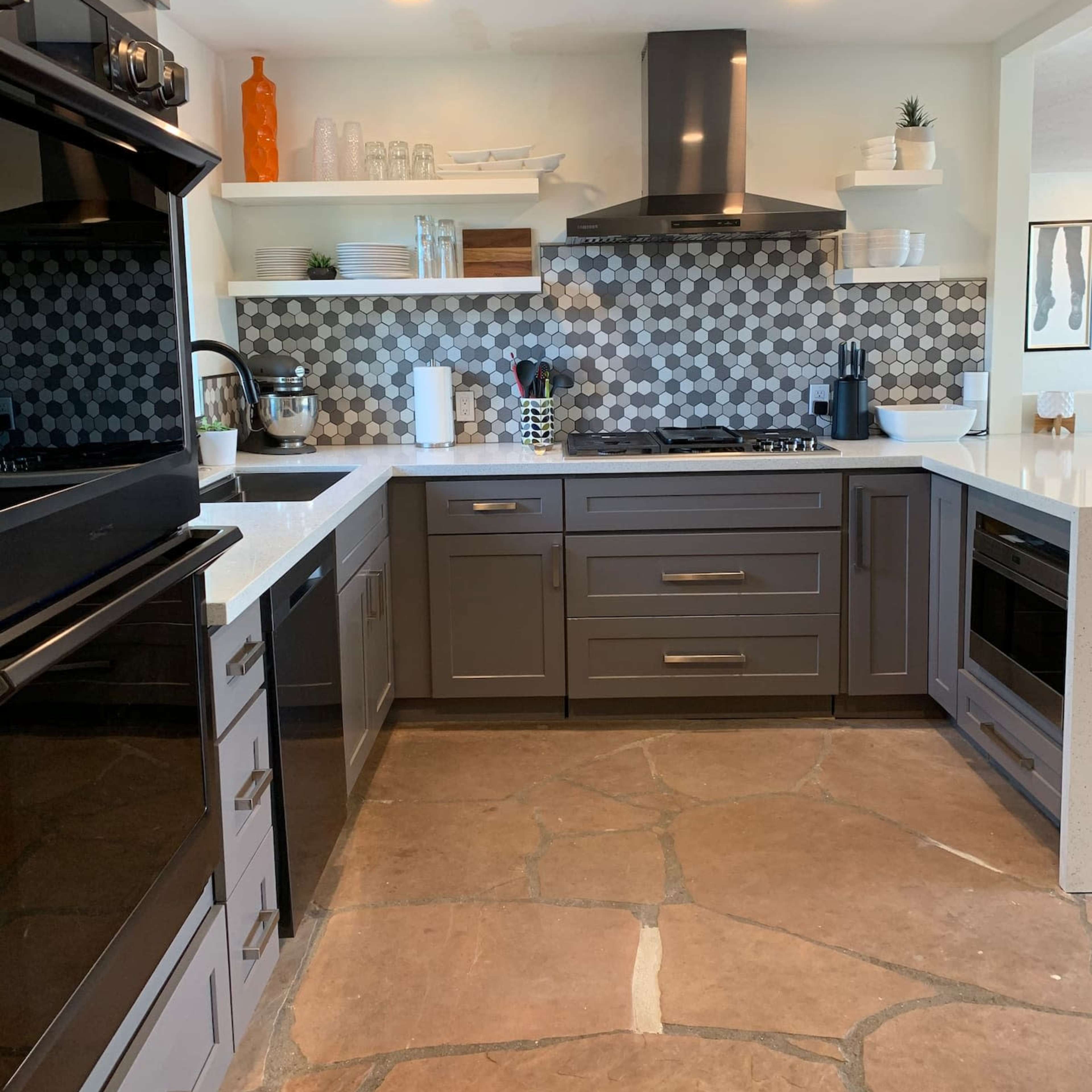 A modern kitchen featuring dark cabinetry, a gas stove, and a patterned tile backsplash, with stone flooring and open shelving displaying various kitchen items.