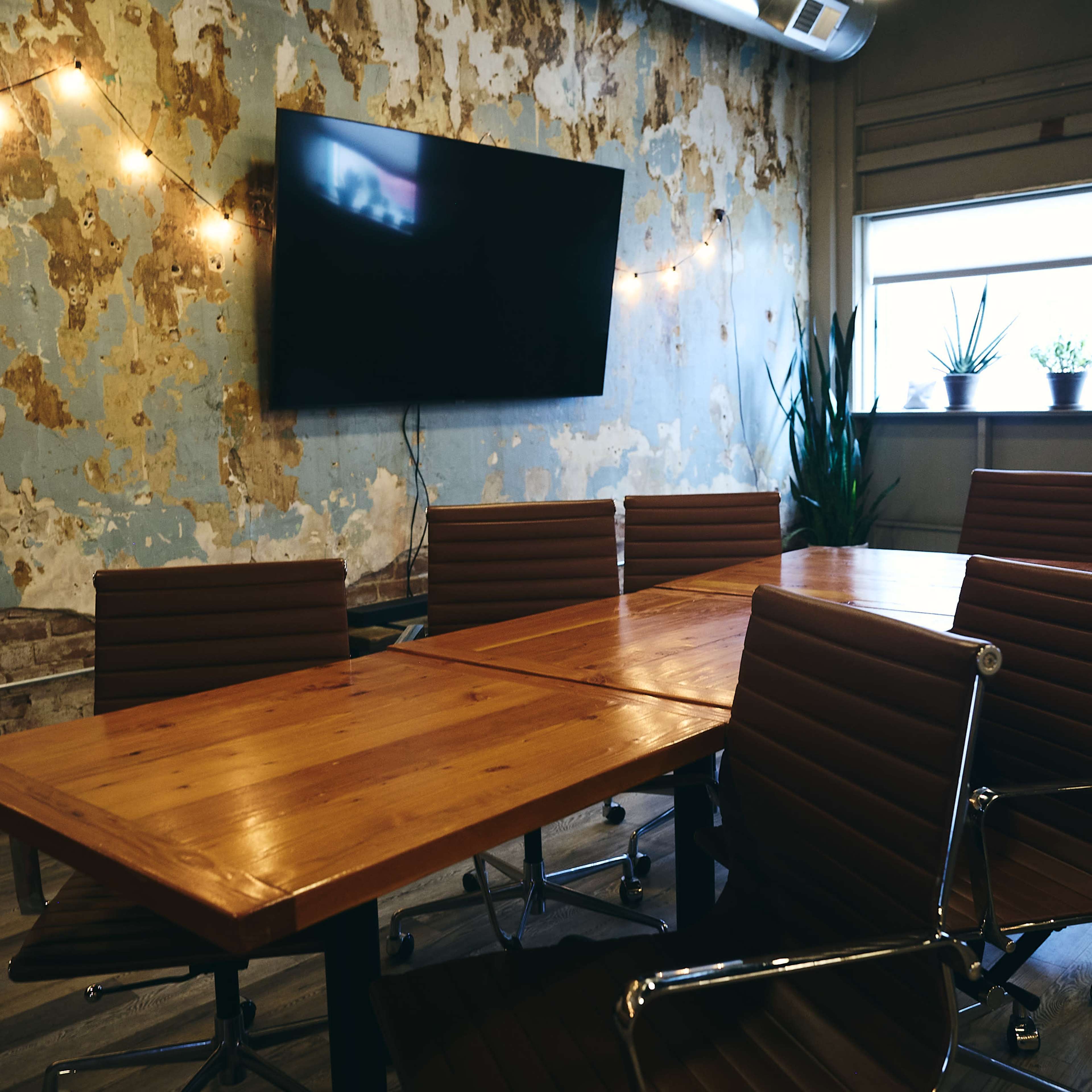 A conference room features a long wooden table surrounded by brown leather chairs, with a large TV mounted on a weathered blue wall and plants visible in the window.