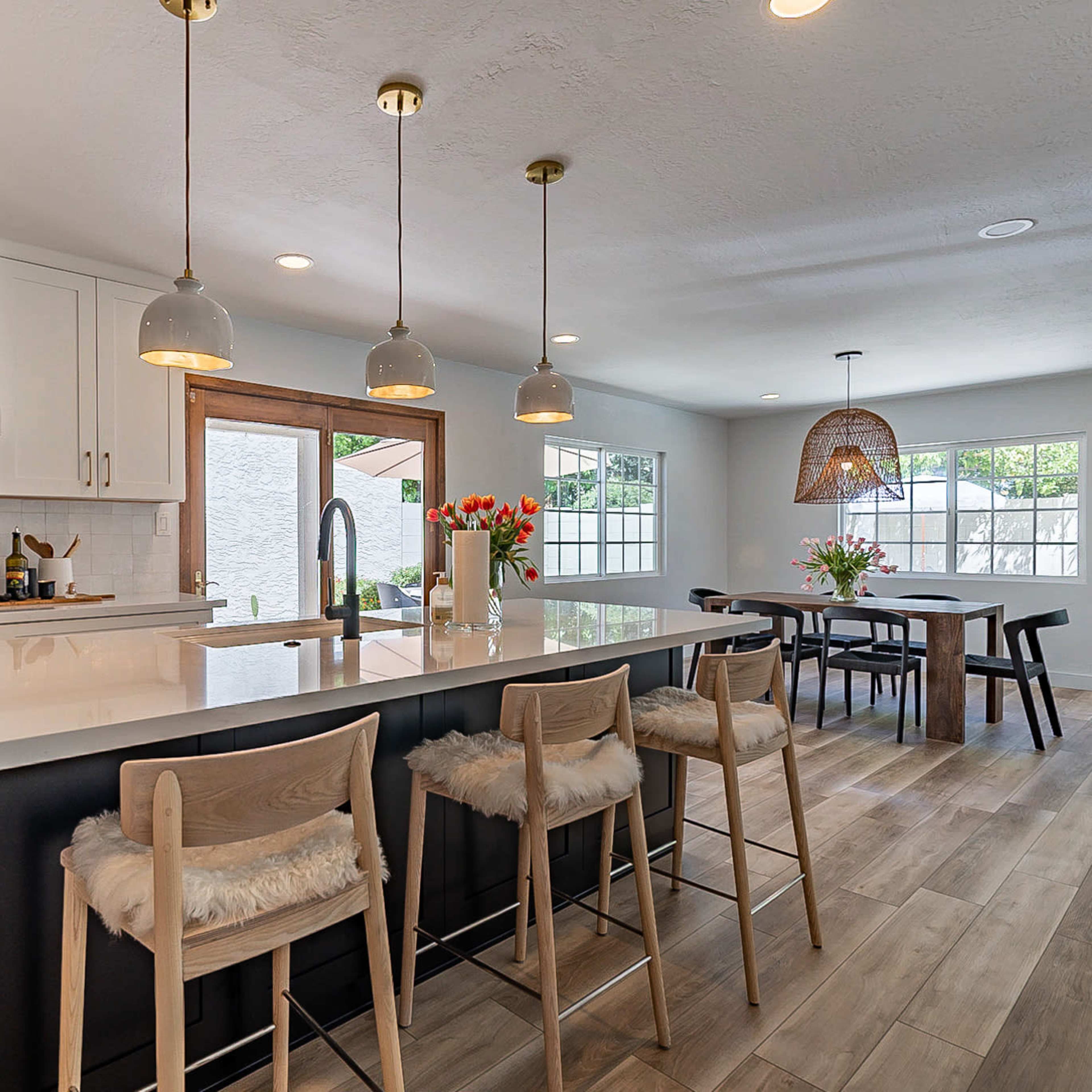 A modern kitchen features a large island with seating, pendant lights, and a dining area with a table and flower arrangements.