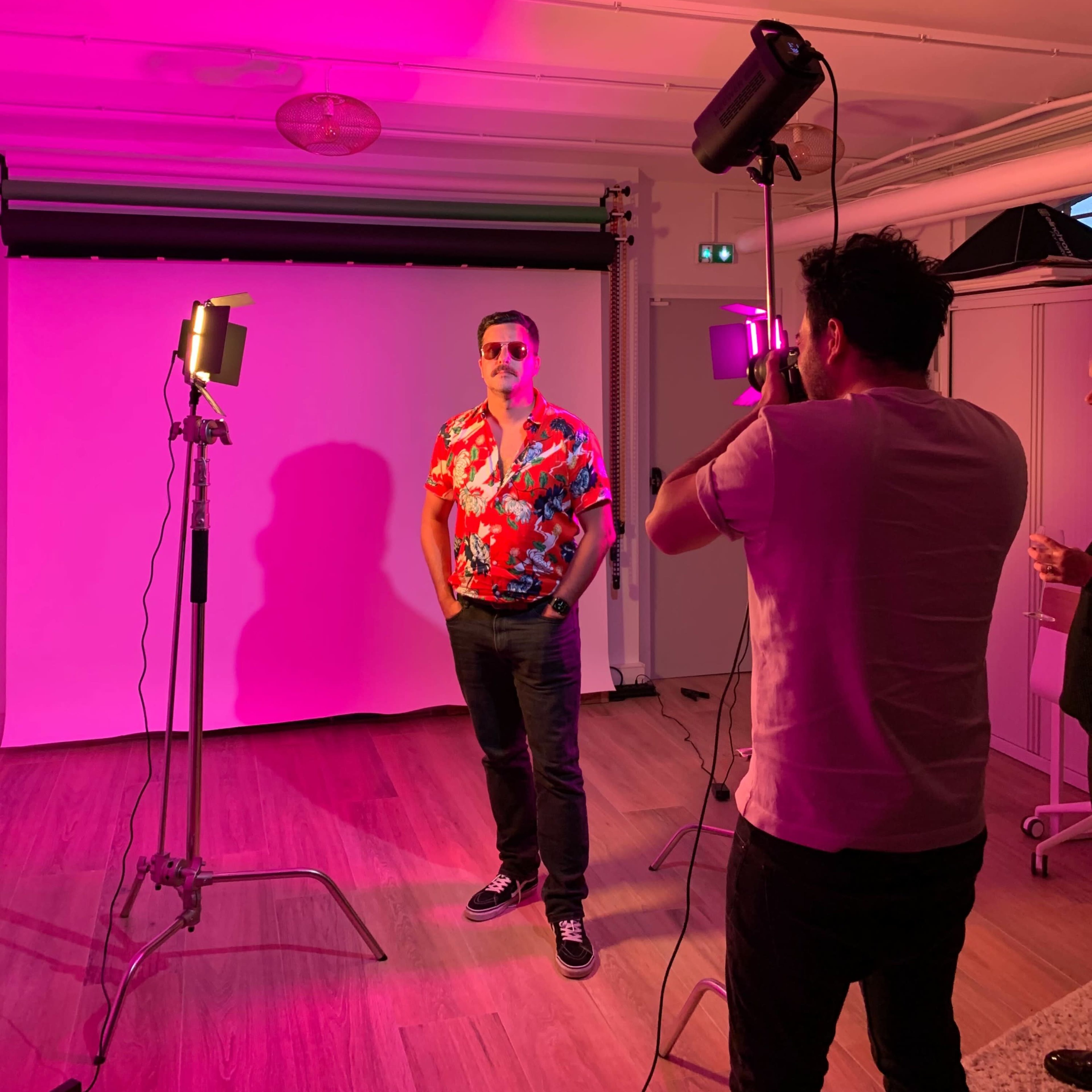 A man in a floral shirt poses for a photo while another person adjusts the camera under pink lighting.