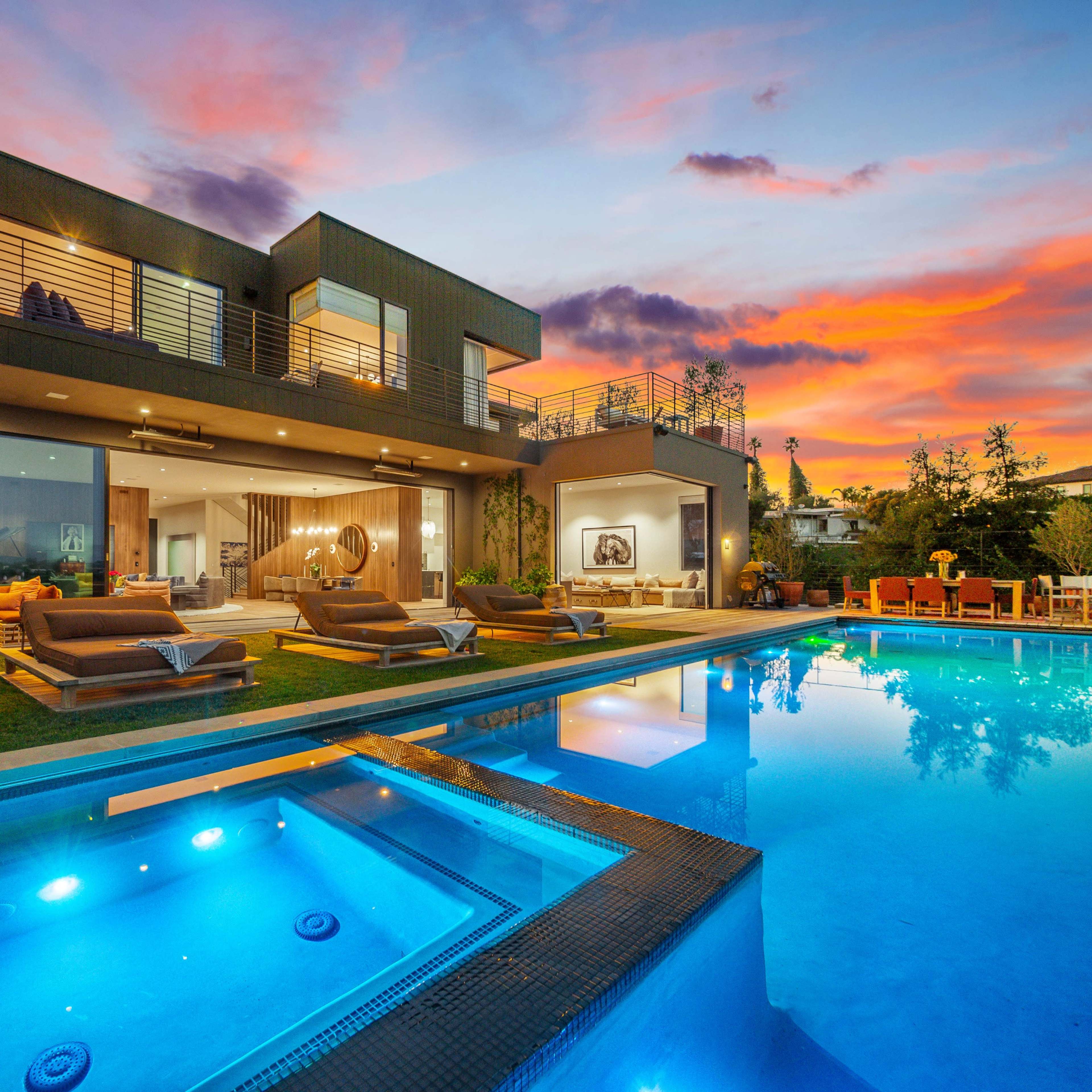 A modern two-story house with large glass windows and a pool in front reflects the colorful sunset sky.