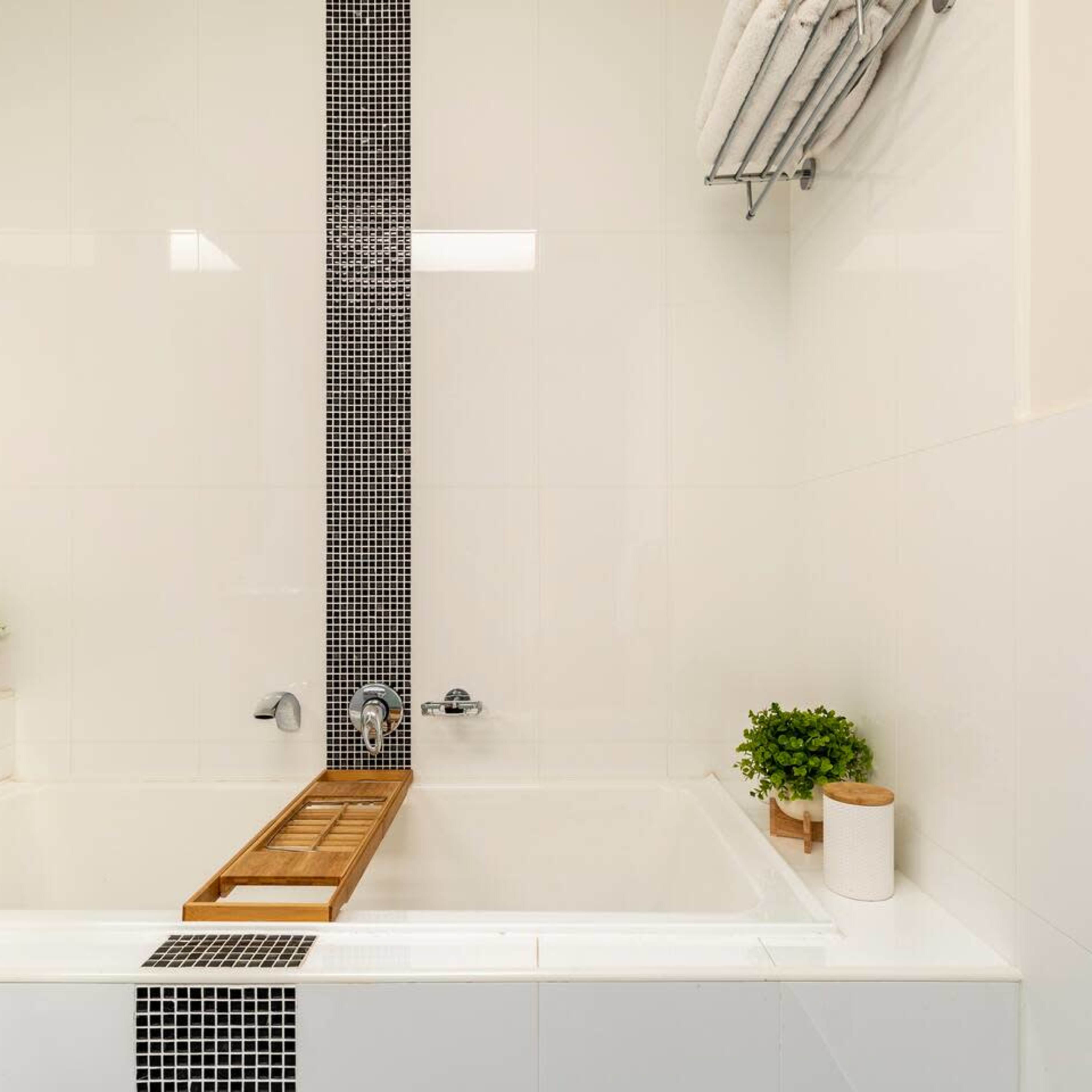 A modern bathroom features a white bathtub with a wooden bath tray, plants, and a black and white tiled wall.