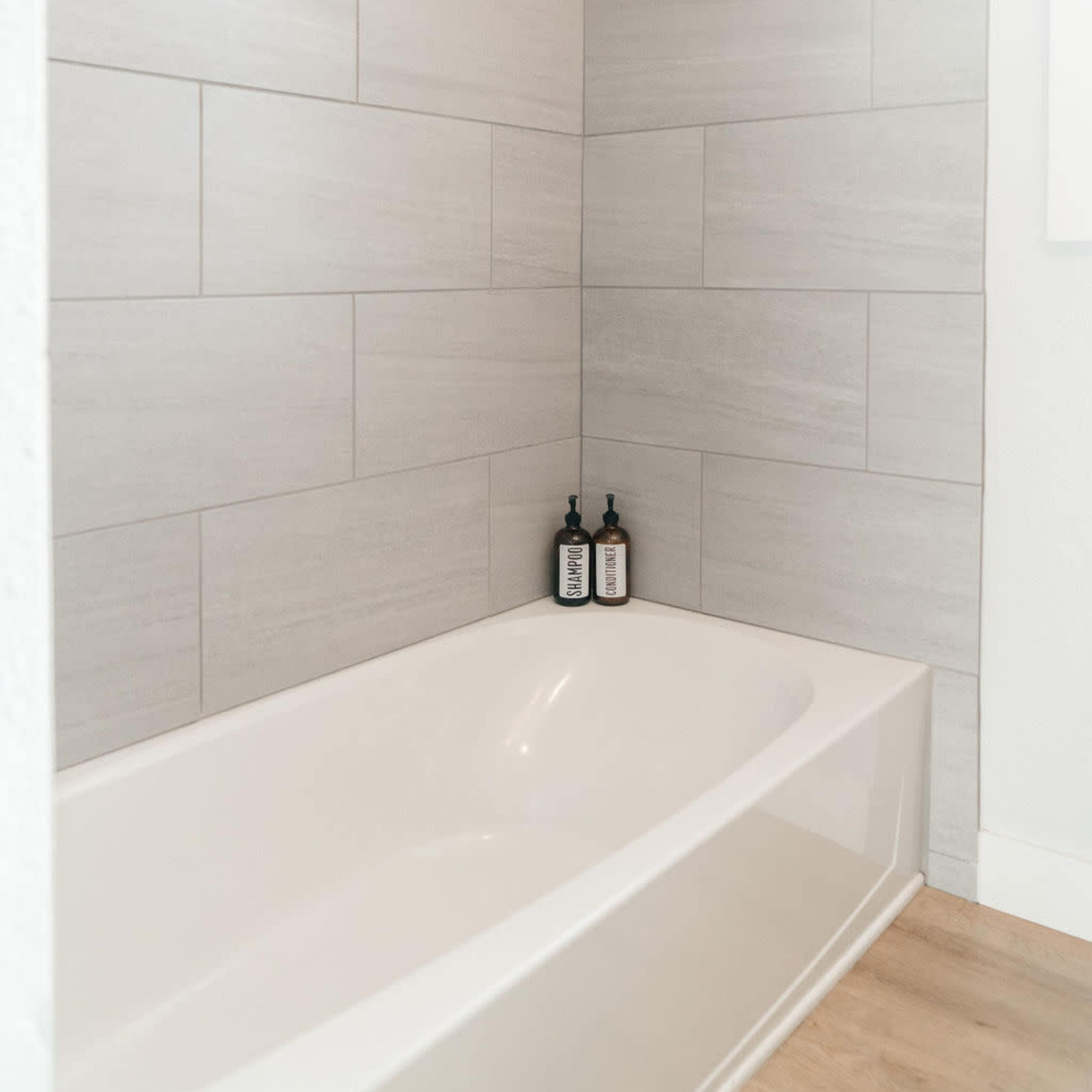 A minimalist bathroom featuring a white bathtub next to light gray tiled walls and two bottles on the edge of the tub.