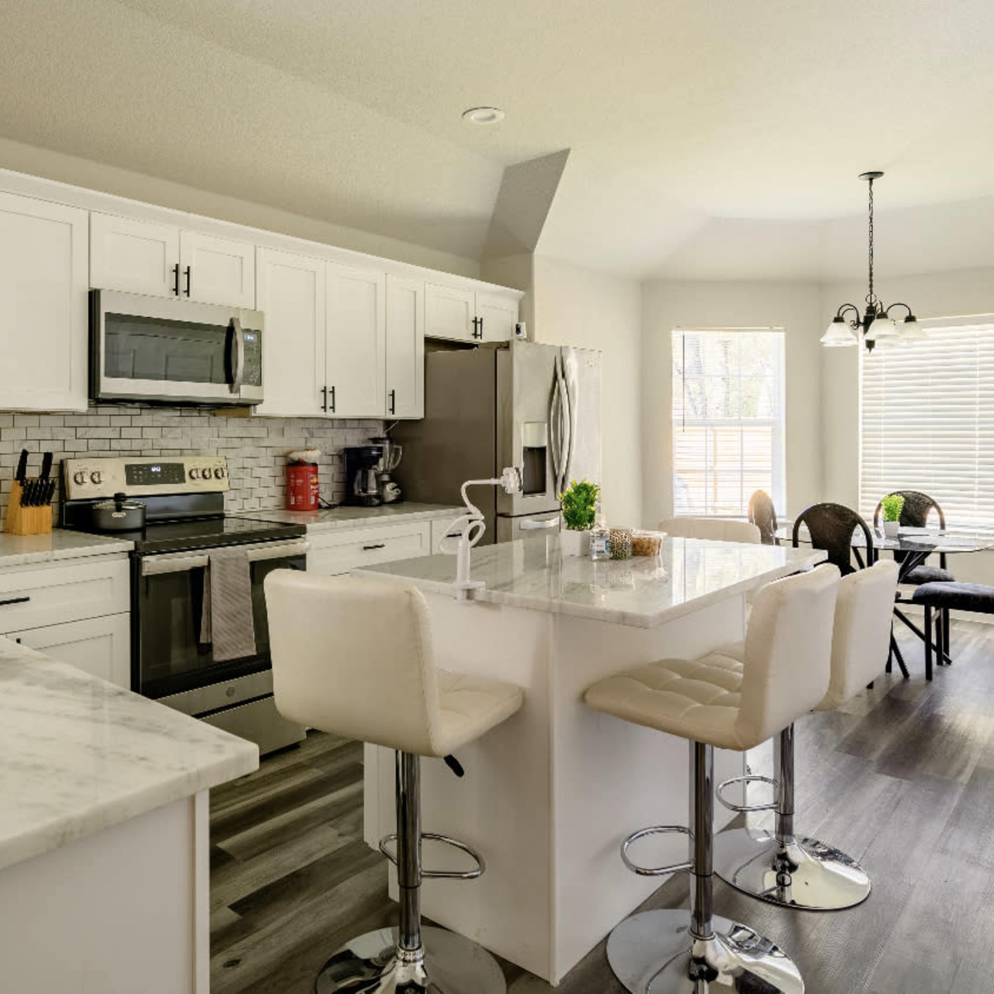 A modern kitchen featuring white cabinets, stainless steel appliances, a marble countertop island with seating, and a dining area with a round table and chairs.
