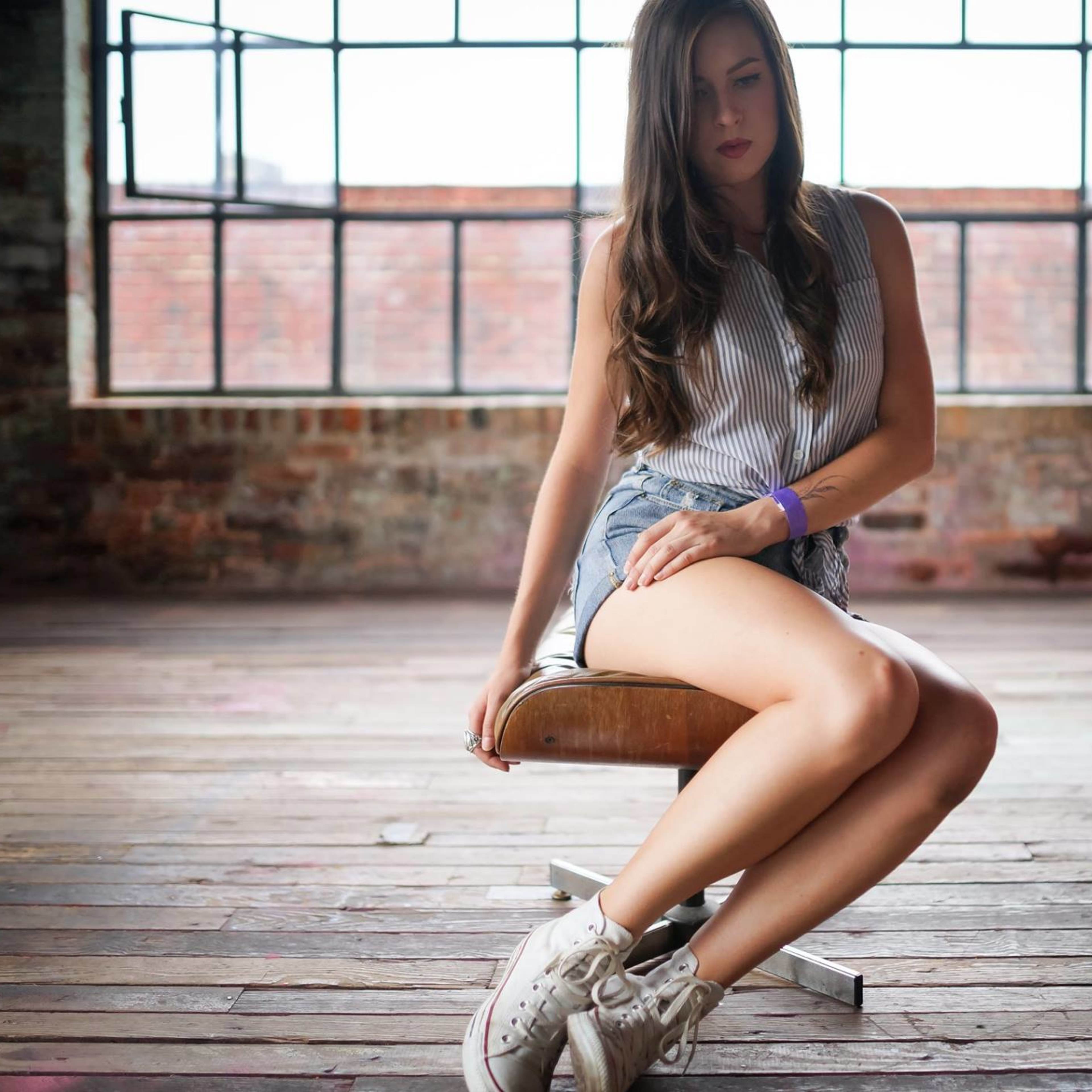 A woman sits on a wooden stool in a spacious, industrial-style room with large windows.