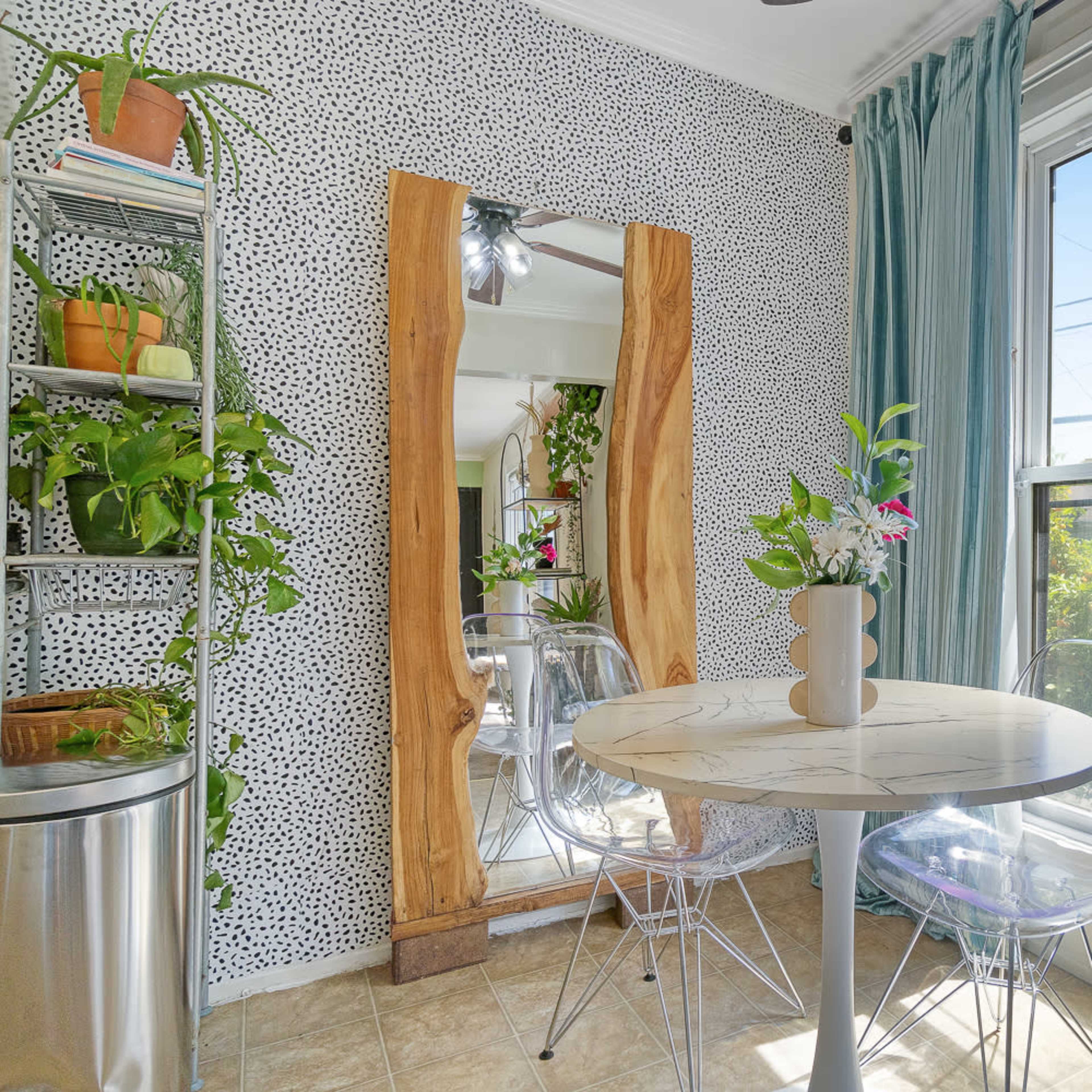 A small dining area features a round marble-top table surrounded by clear plastic chairs, with green plants and a large wooden mirror against a speckled wall.