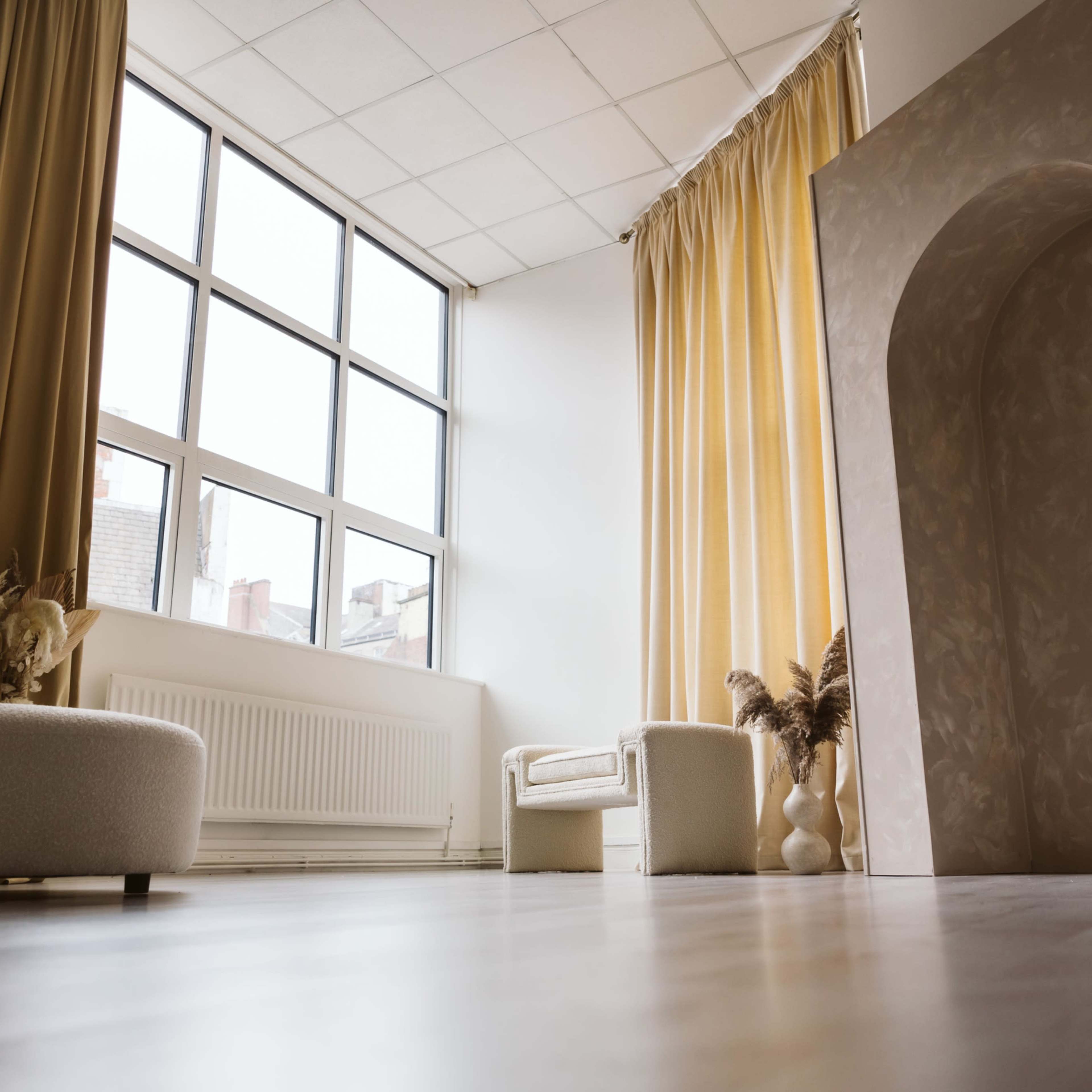 A spacious room with large windows, a light-colored archway, and minimal furniture including a sofa and an armchair.
