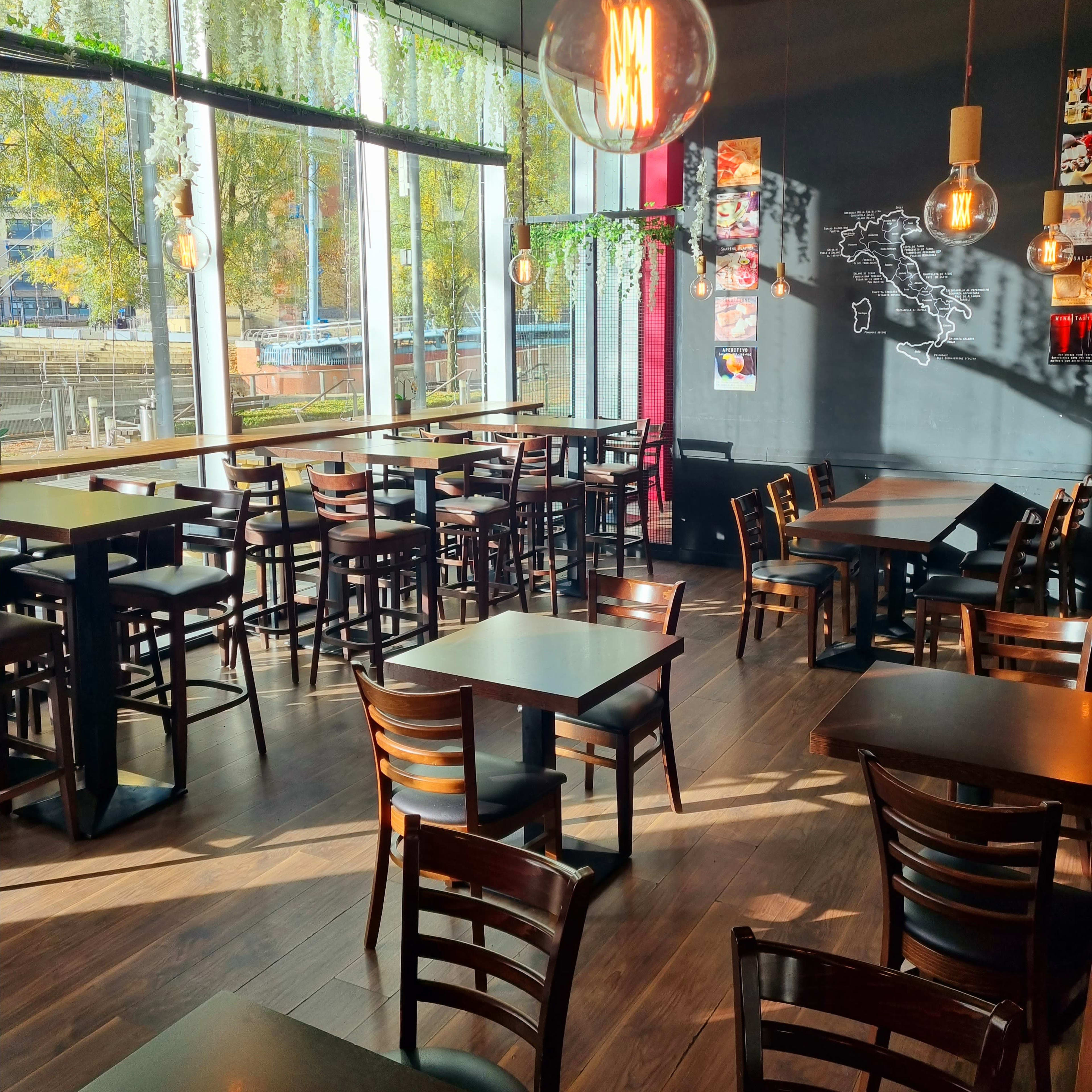 A restaurant interior with wooden tables and chairs, large windows allowing natural light, and hanging pendant lights.