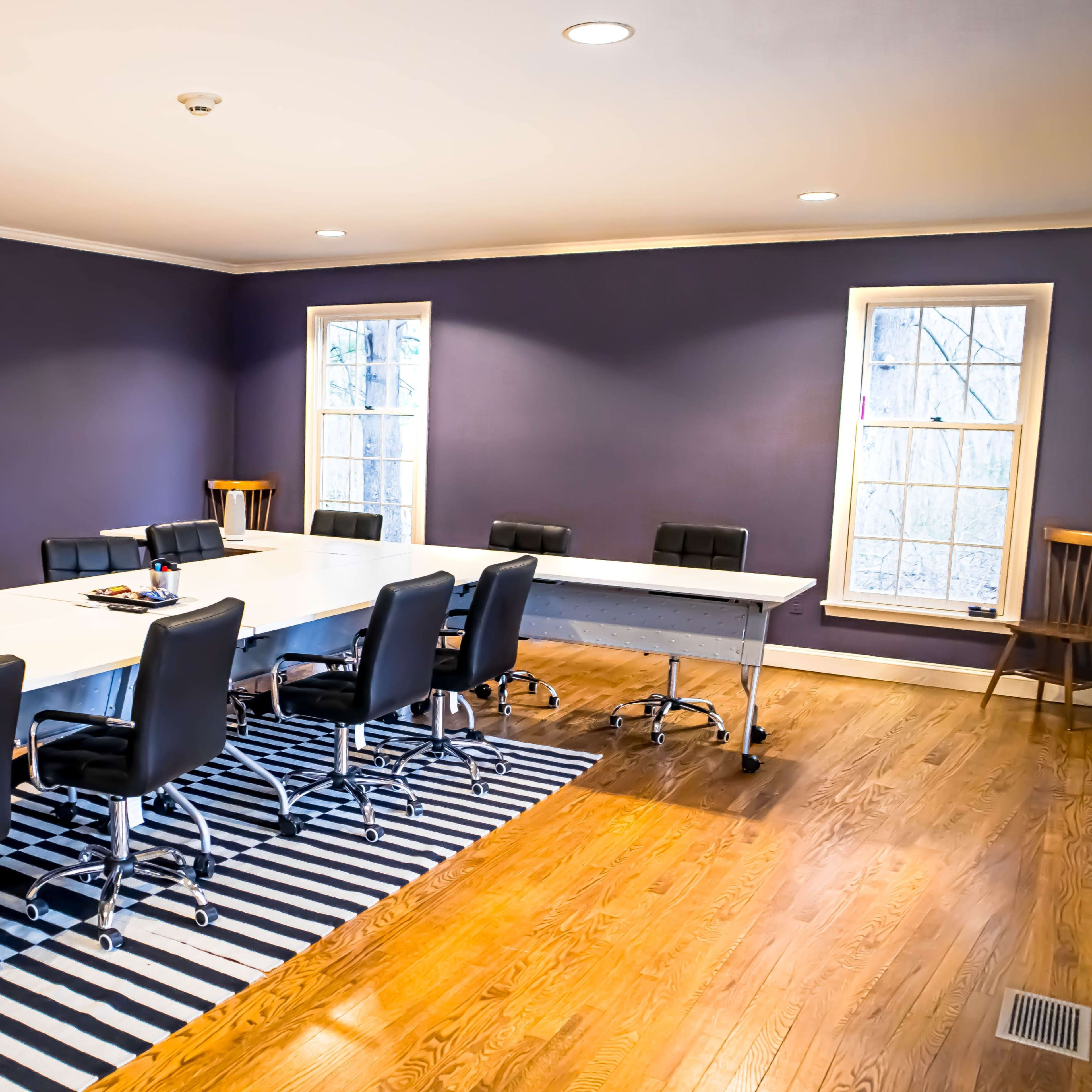 A spacious conference room features a long white table surrounded by black chairs on a striped rug with windows letting in natural light.