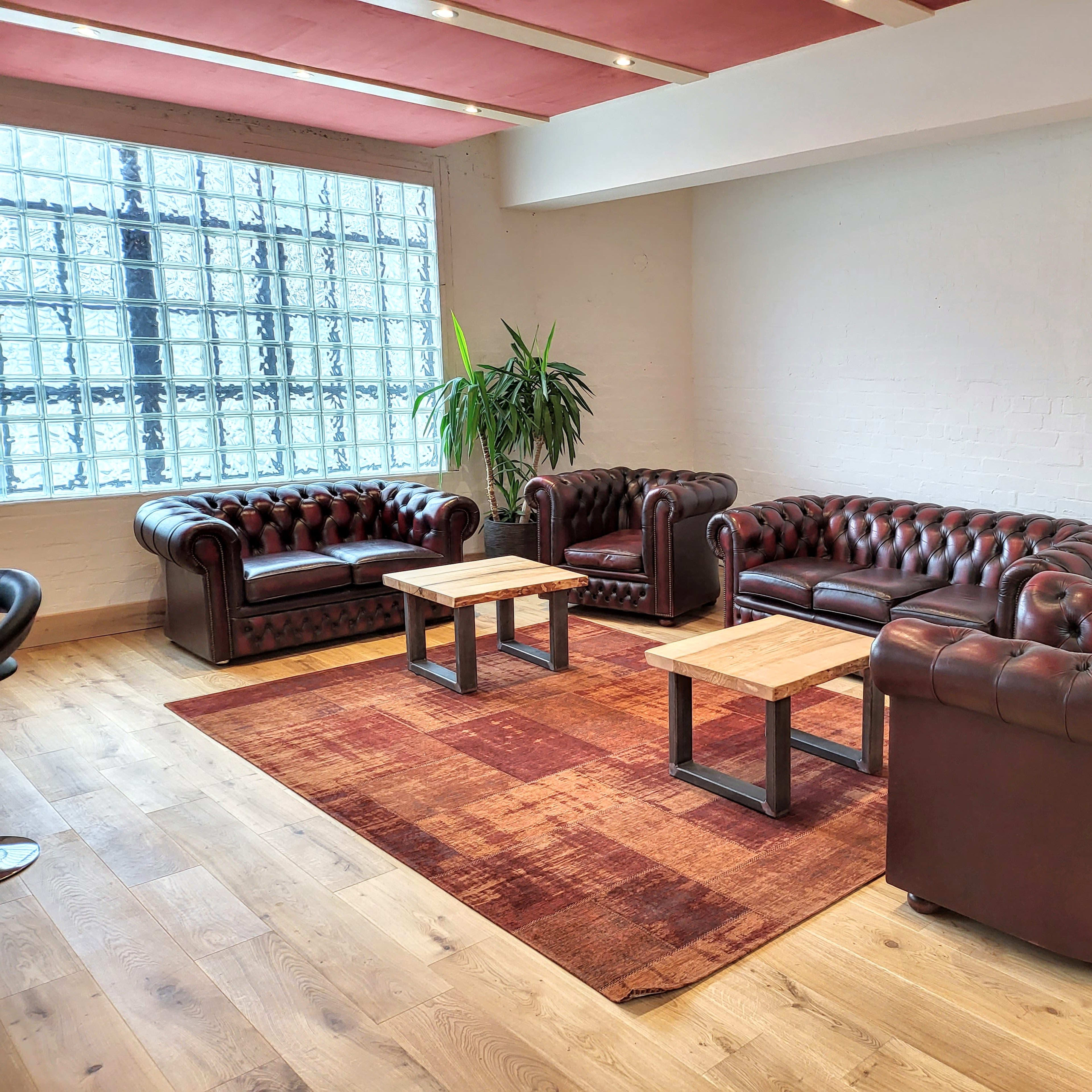 A room with four brown leather sofas arranged around two wooden tables on a carpet, with a large glass block window and a potted plant in the background.