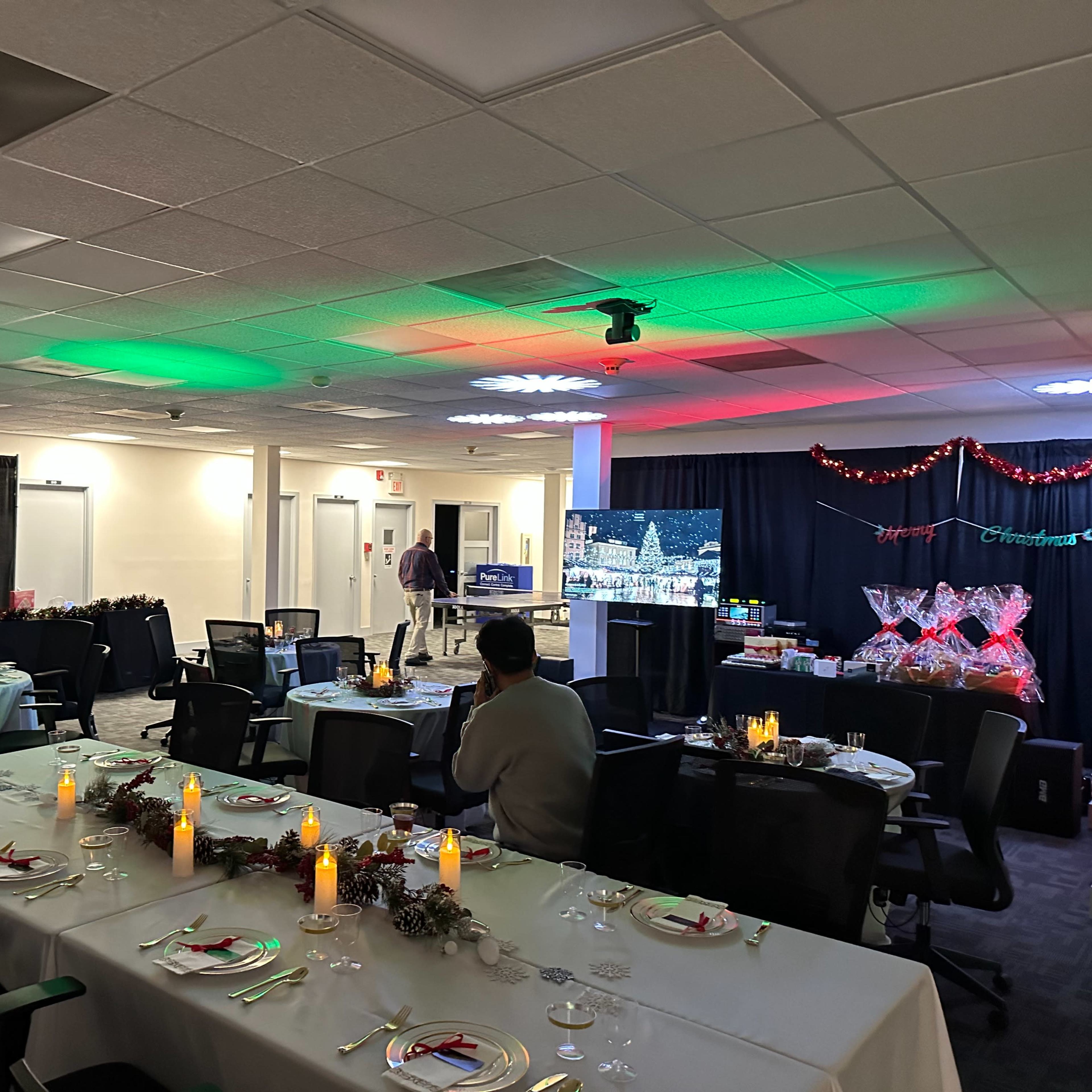 A spacious room is set up for a festive gathering, featuring decorated tables with candles and a backdrop of holiday-themed decorations.