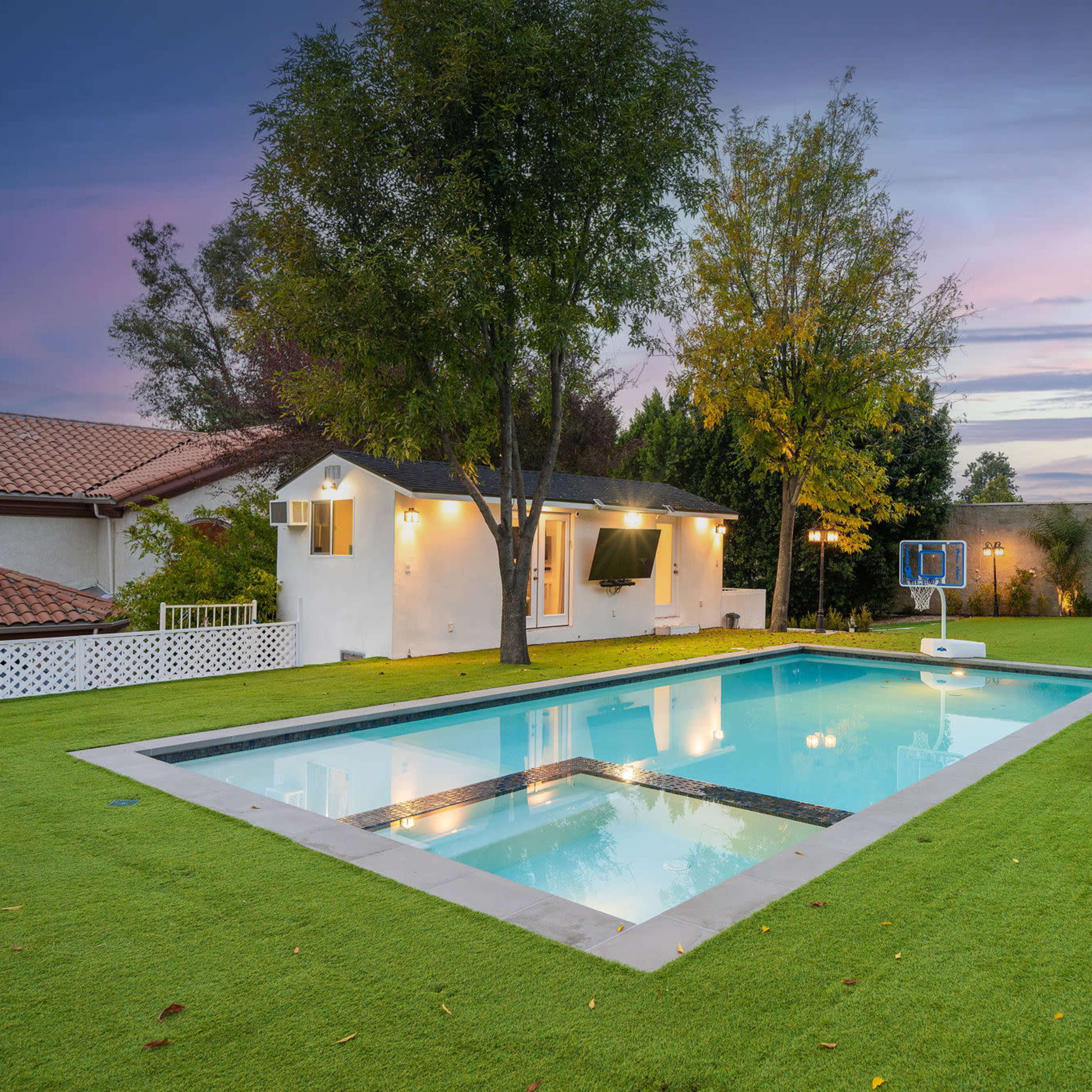 A backyard featuring a rectangular swimming pool surrounded by lush green grass and two lounge chairs, with a small white building and trees in the background under a twilight sky.
