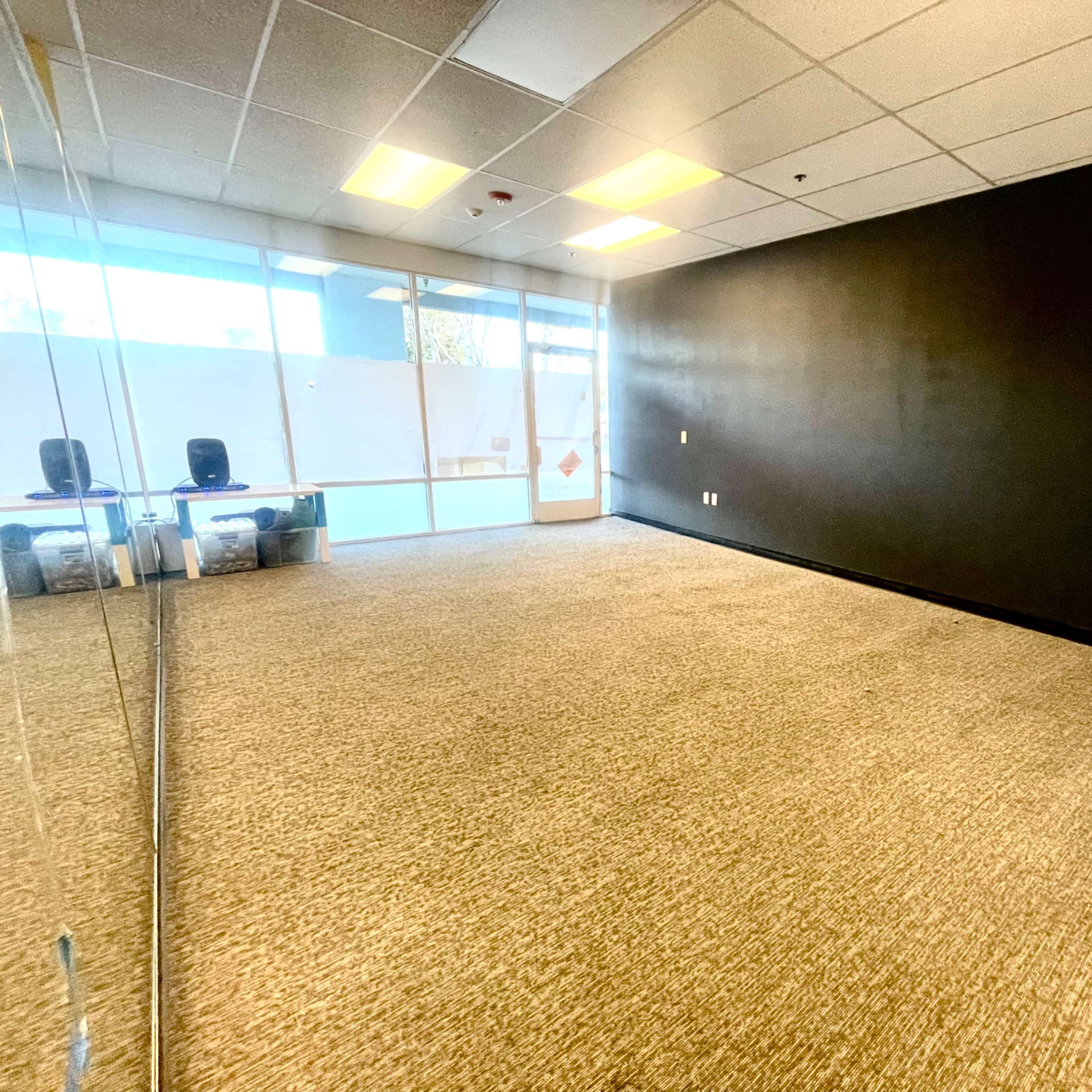 A spacious, empty room with a dark wall and a large mirror on one side, featuring a carpeted floor and window light coming from the entrance.