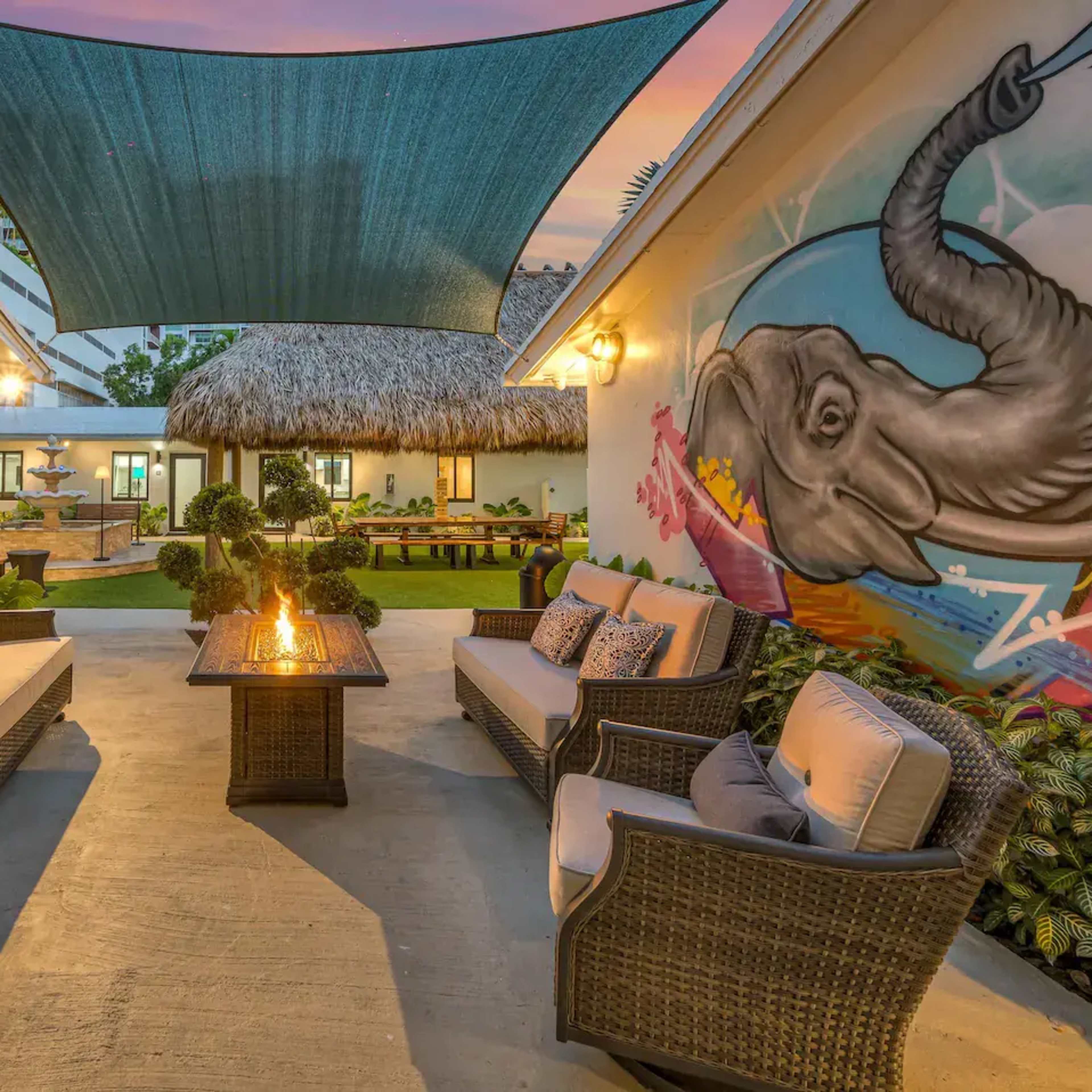 An outdoor seating area with wicker furniture surrounded by vibrant murals of a tiger and an elephant, complemented by a shaded canopy and a fire pit.