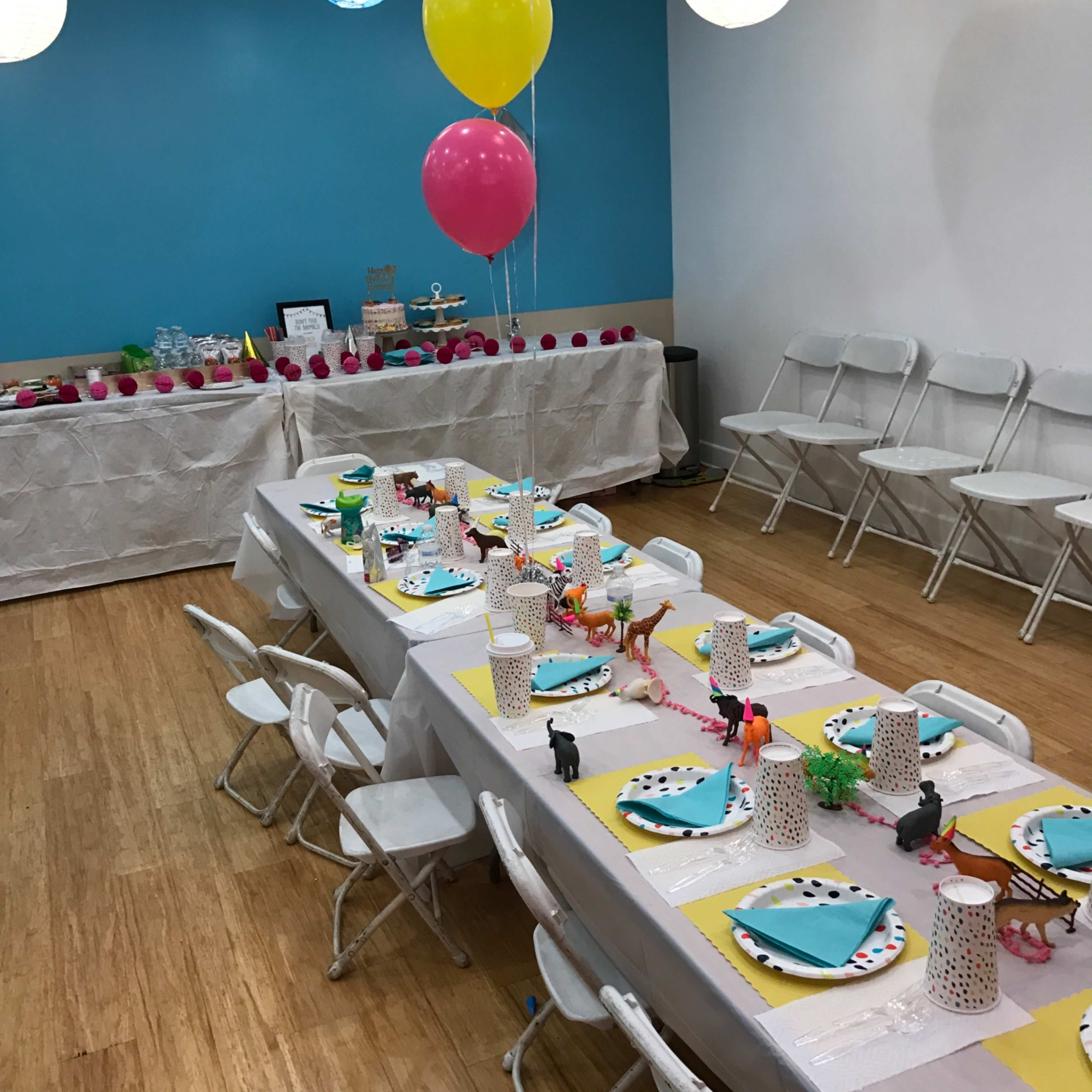 A decorated party room features a long table set with colorful plates, animal-themed centerpieces, and balloons, surrounded by white folding chairs.