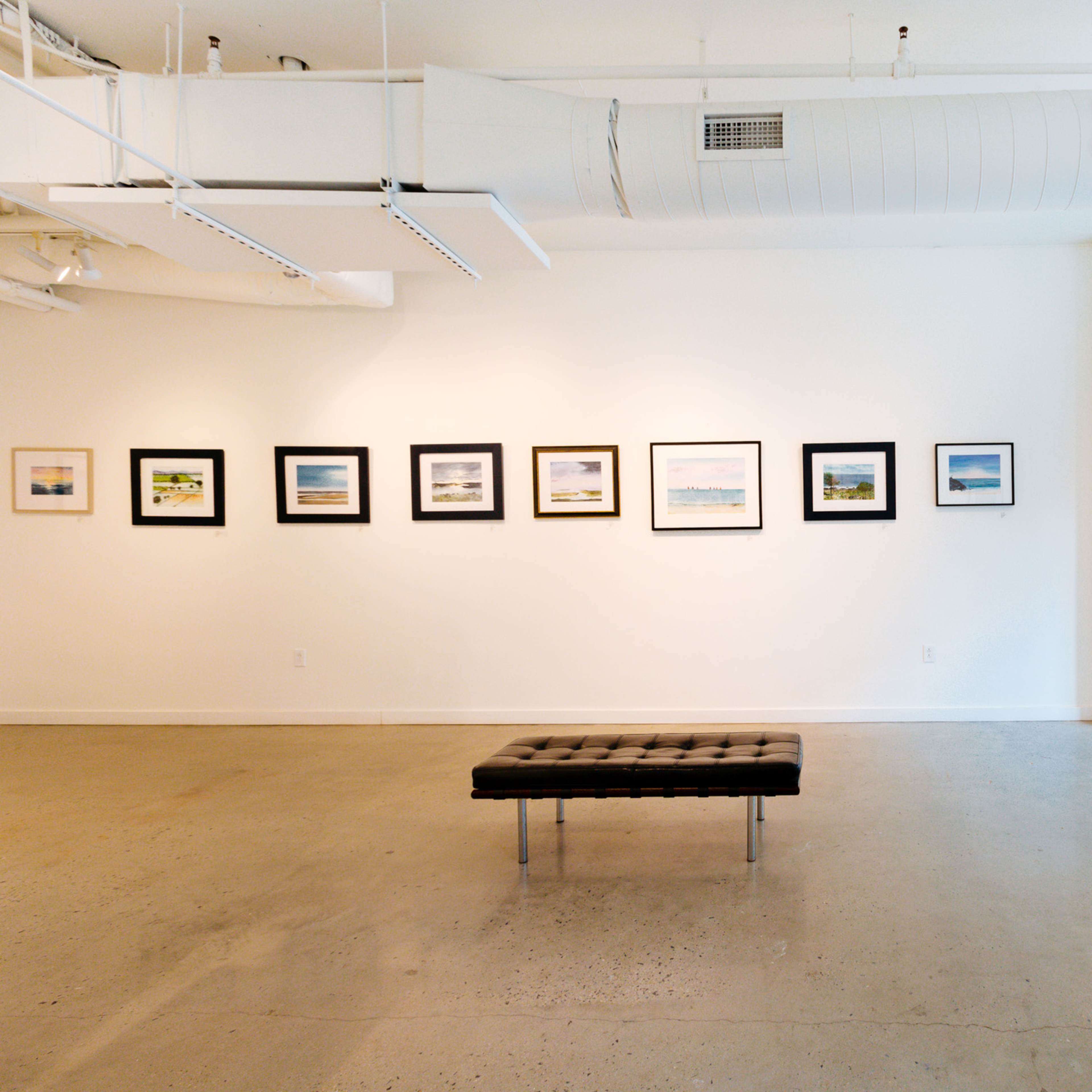 An art gallery with several framed paintings displayed on the walls and a black bench in the center of the room.