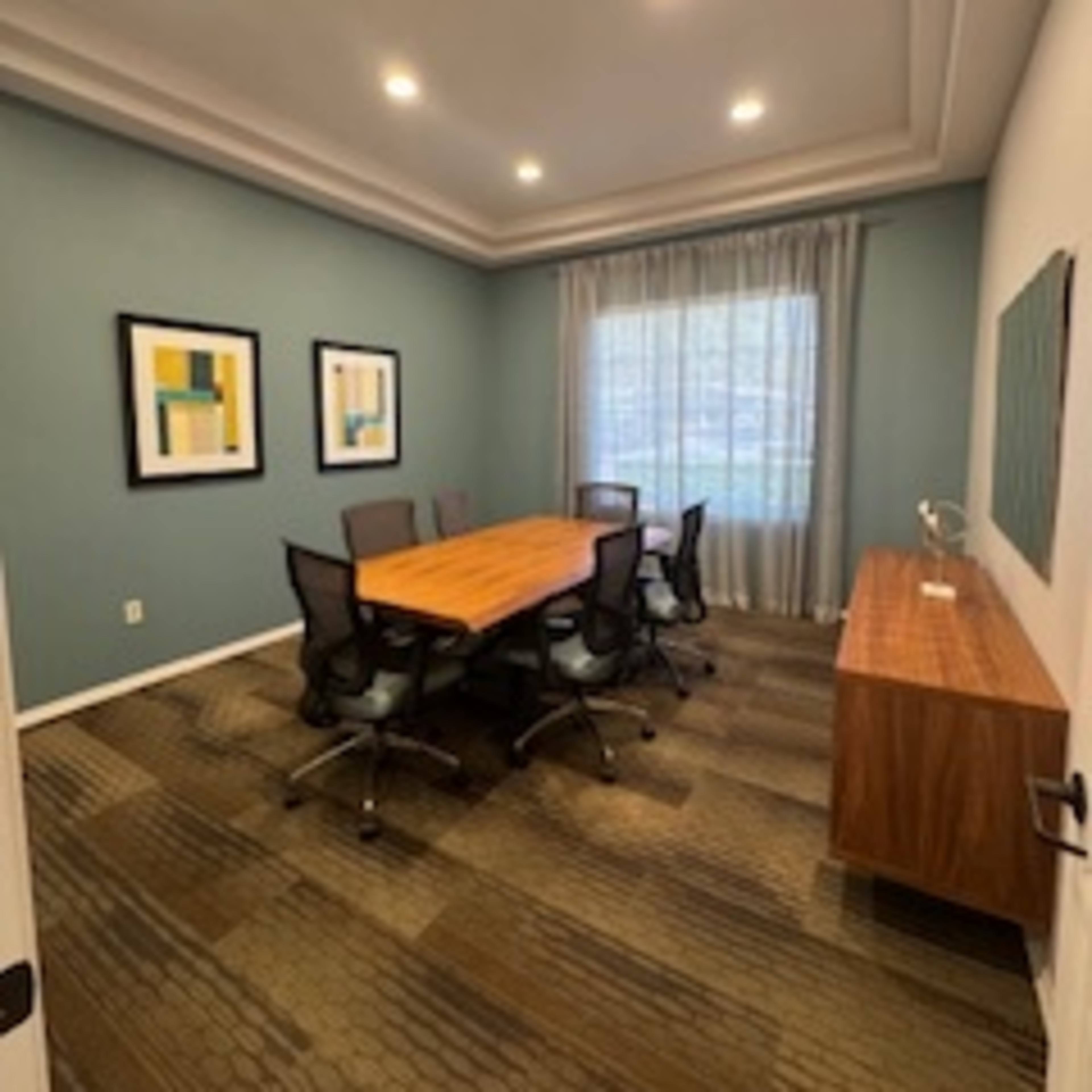 A conference room with a wooden table surrounded by six chairs, framed artwork on the walls, and a window with sheer curtains.