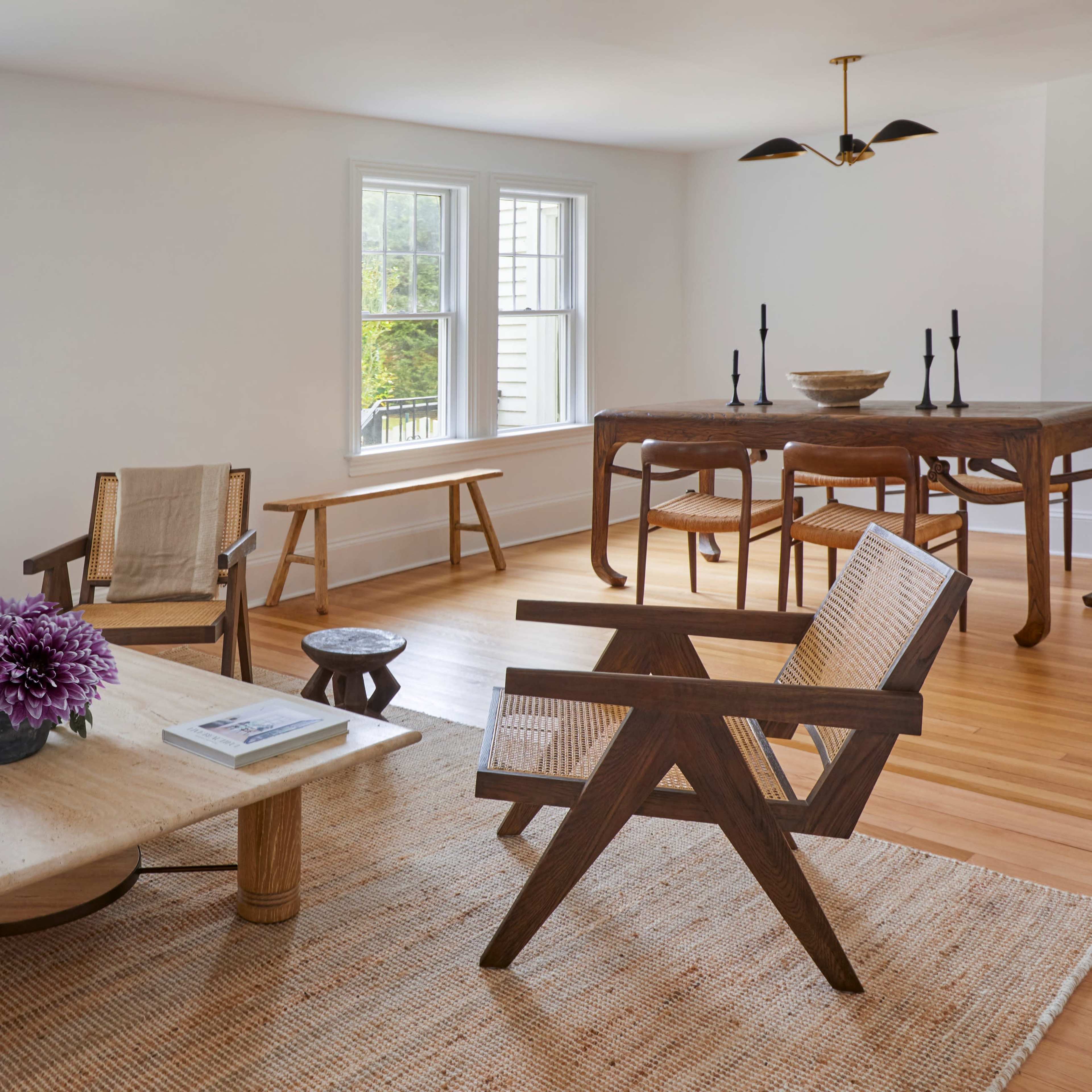 A bright, minimalist living space featuring two wooden chairs, a low coffee table, and a dining table with candles, set against a backdrop of windows and wooden flooring.