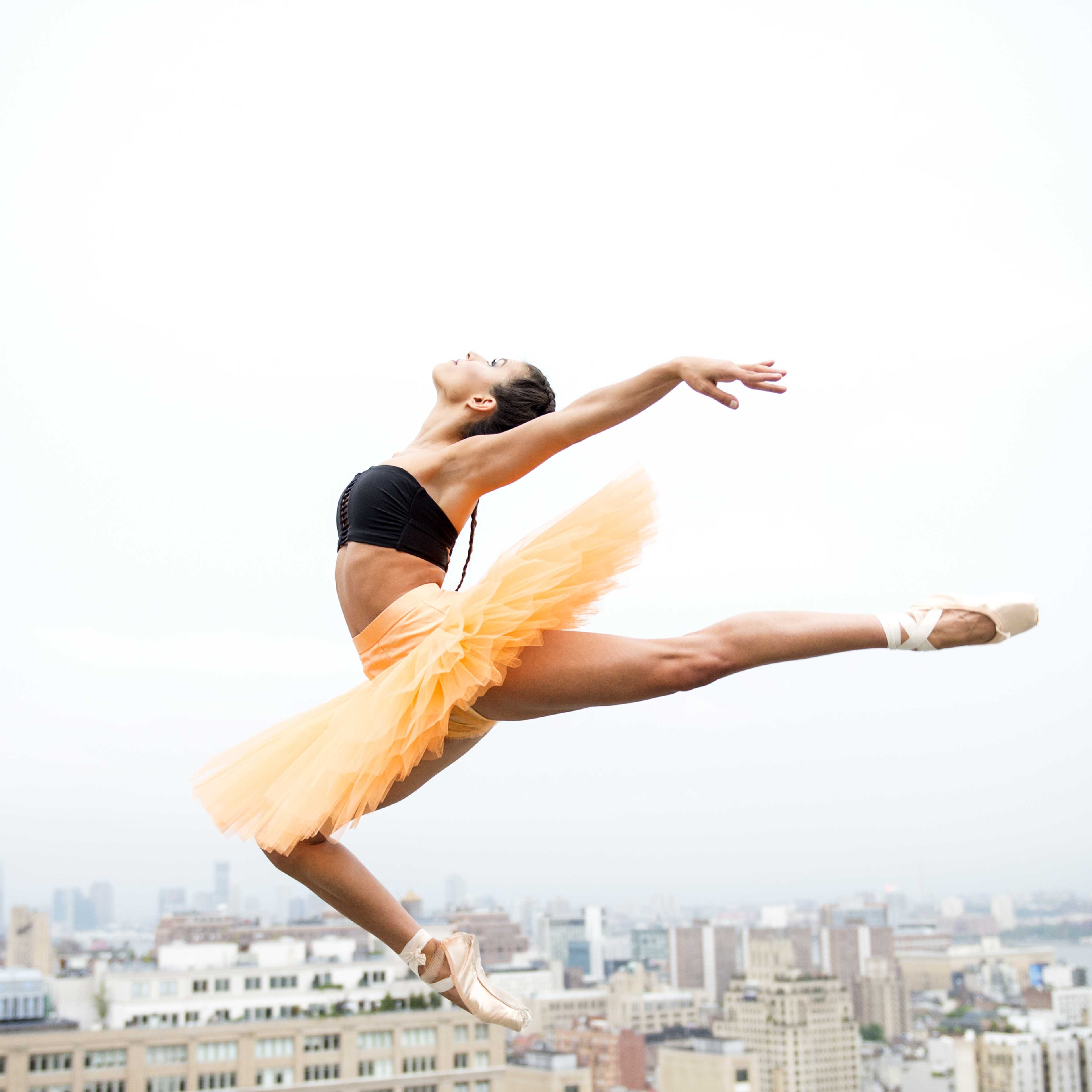 A ballet dancer in an orange tutu performs an aerial leap against a city skyline backdrop.