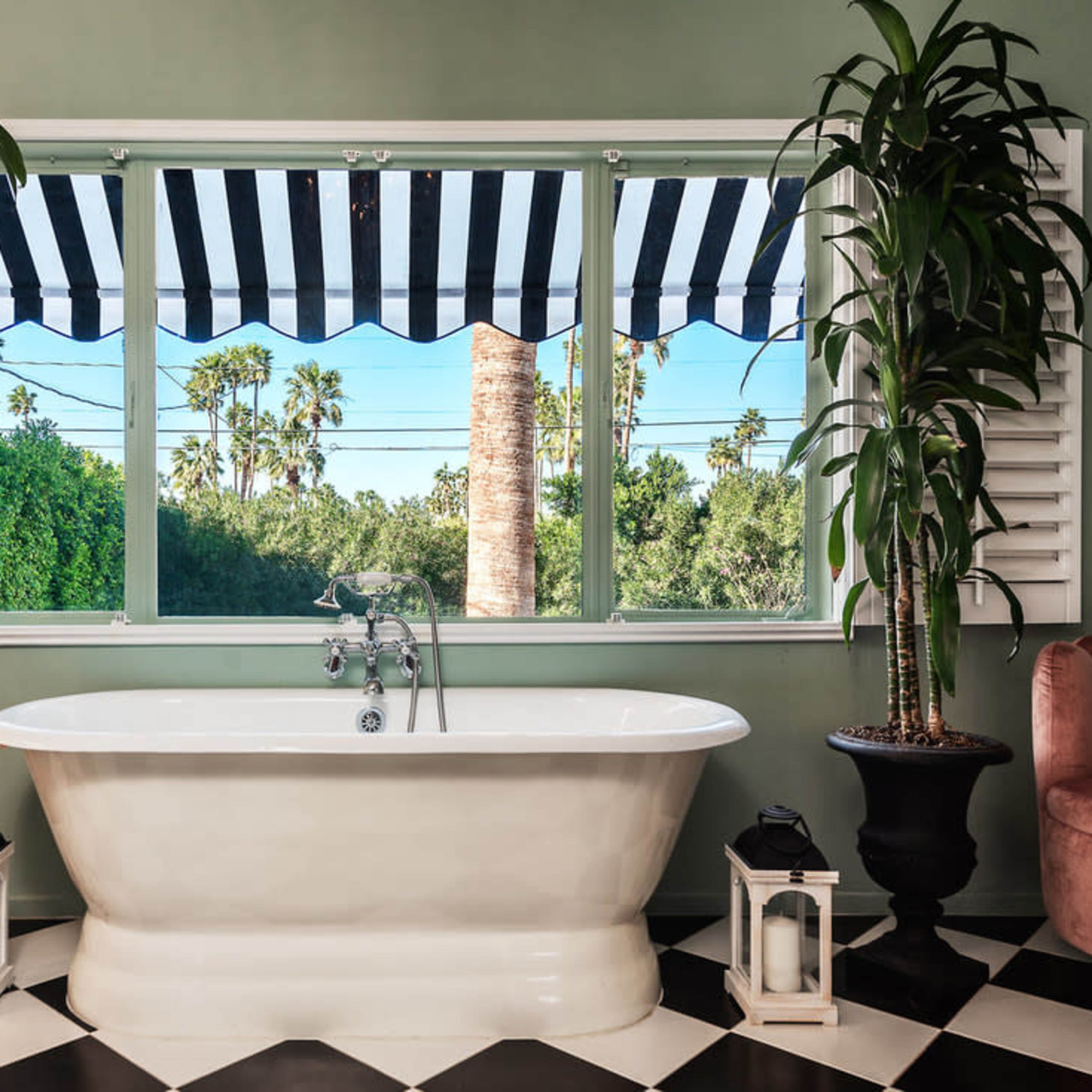 A freestanding white bathtub is positioned between large windows adorned with black and white striped awnings, framed by potted plants, with a checkerboard floor below.