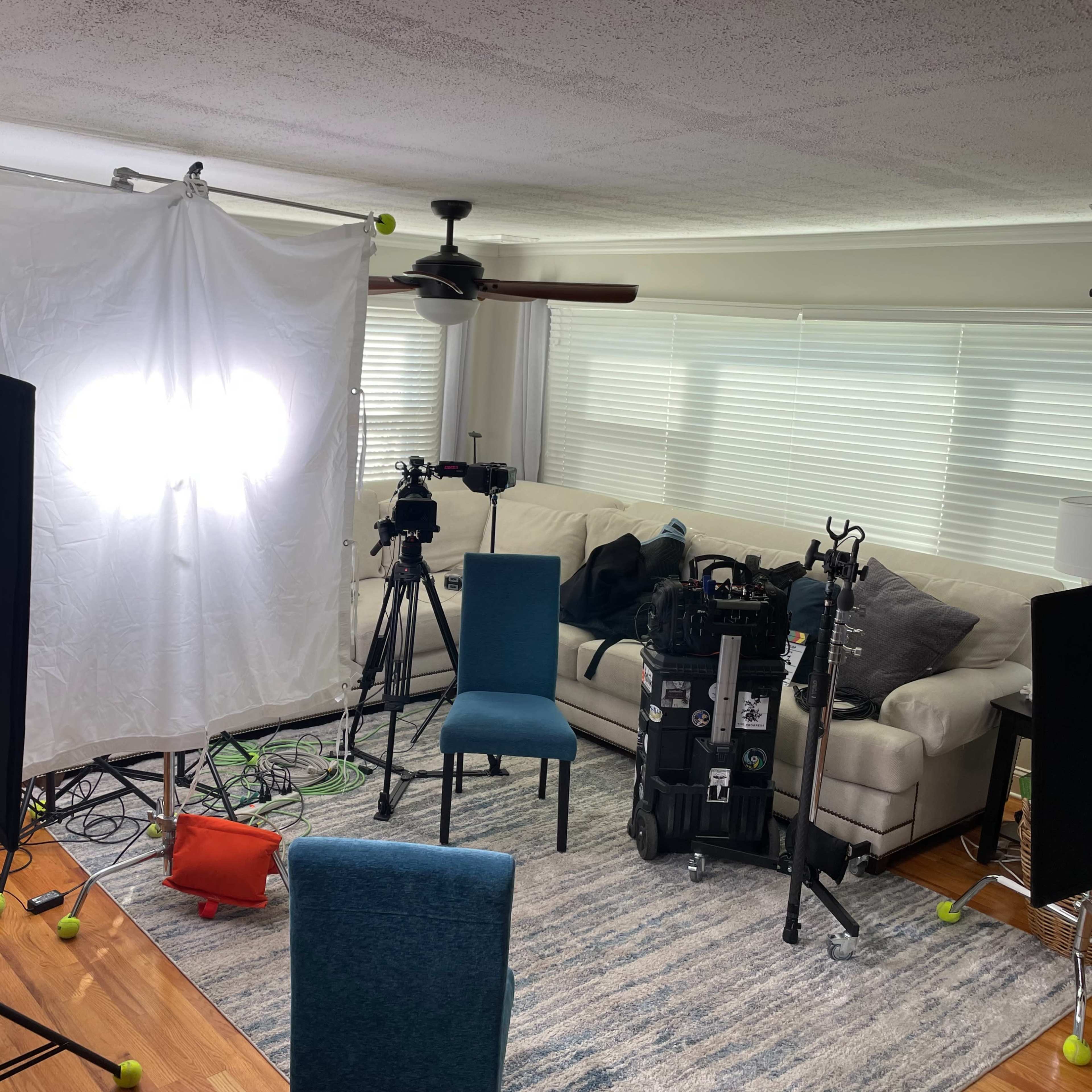 A film set is arranged in a living room, featuring cameras, lighting equipment, and a blue chair on a patterned rug.