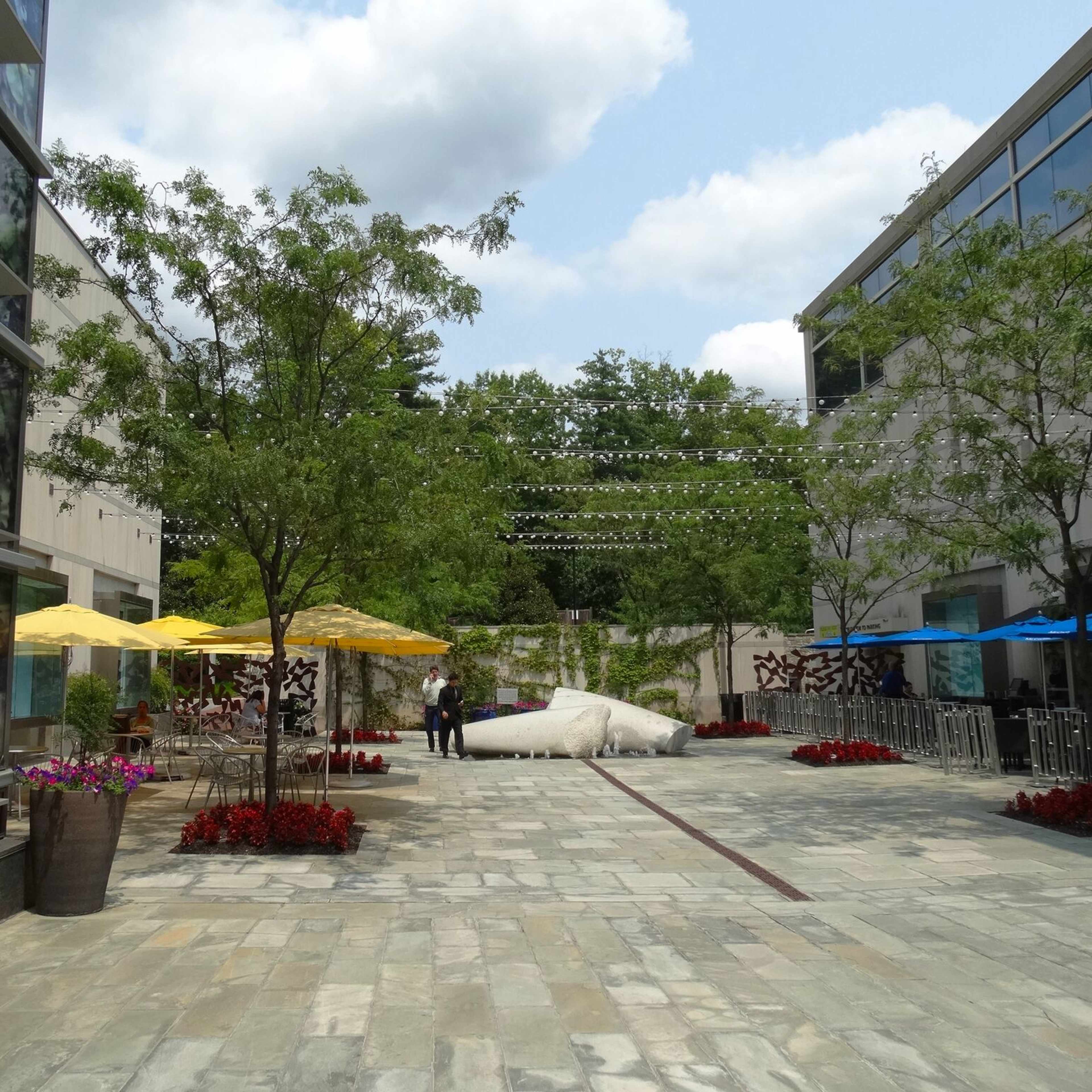 A spacious outdoor area between two buildings, featuring stone paving, colorful umbrellas, trees, and flower beds.