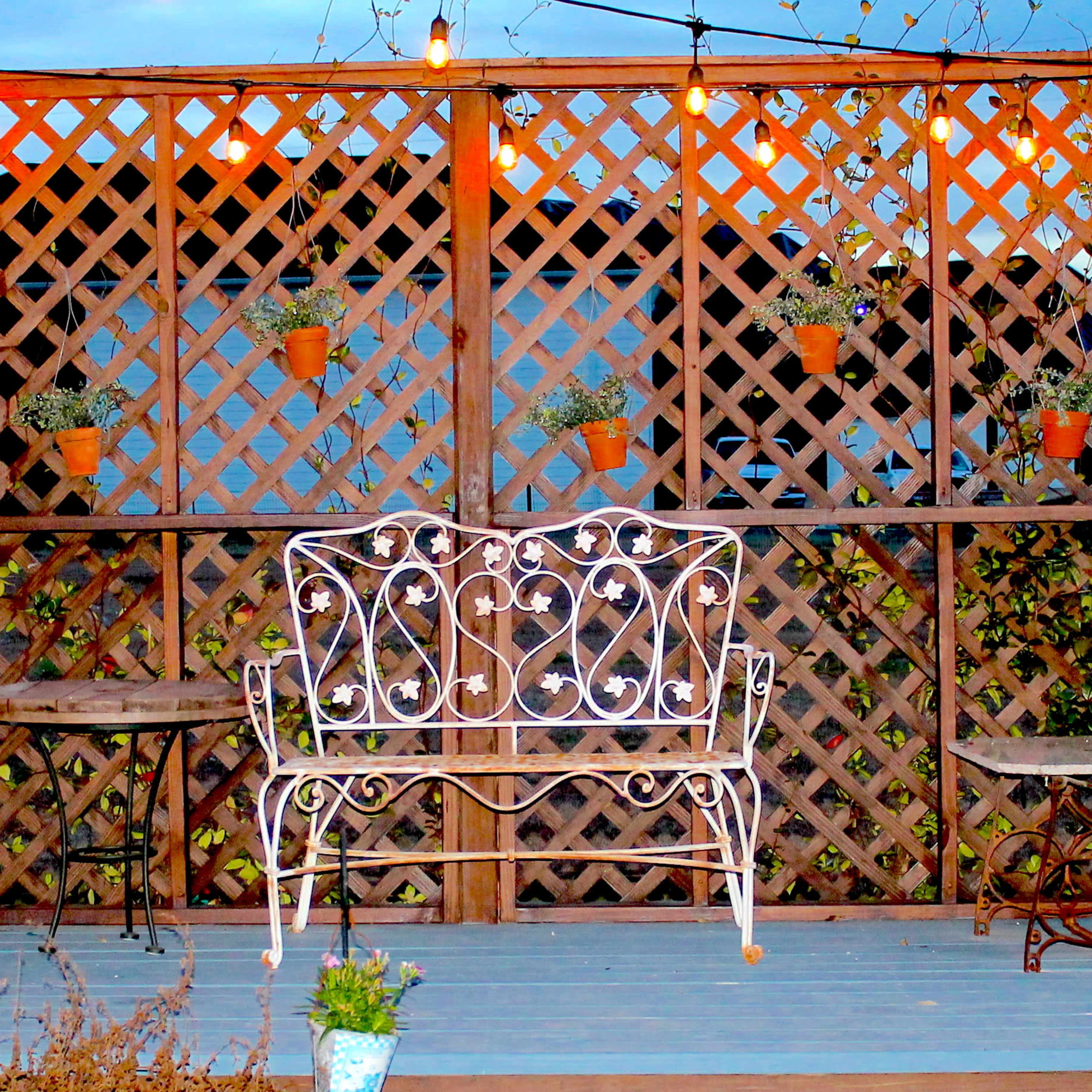 A wooden trellis with hanging potted plants and a white wrought-iron bench in front of two small tables.